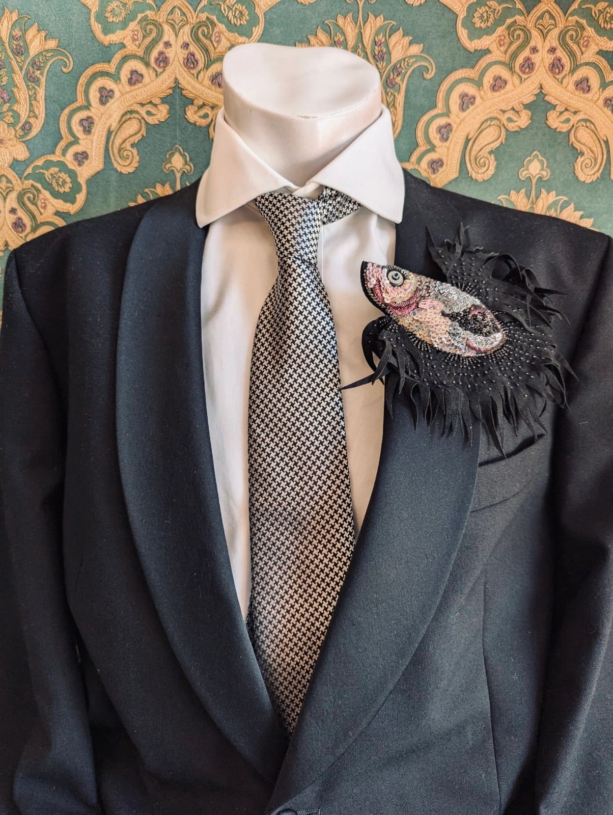 Black suit with a patterned tie and decorative pocket square against an ornate wallpaper background. Pinned to the lapel is the Broochella men's pesce, siamese hand beaded pink, black and silver brooch.