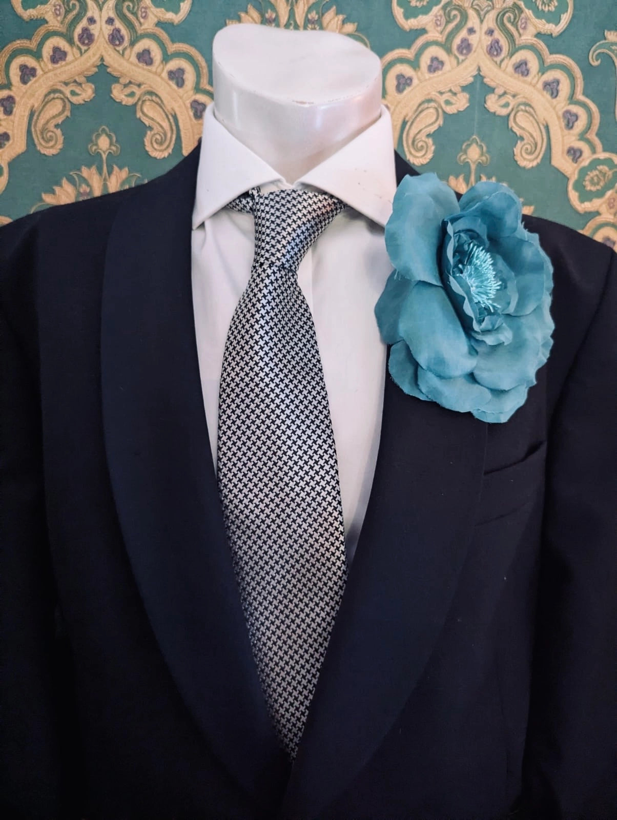 Broochella men's signorina fioritina handmade fabric flower brooch in blue. It is pinned on the label of a black suit jacket that is on a men's mannequin. The background is green and gold damask wallpaper.