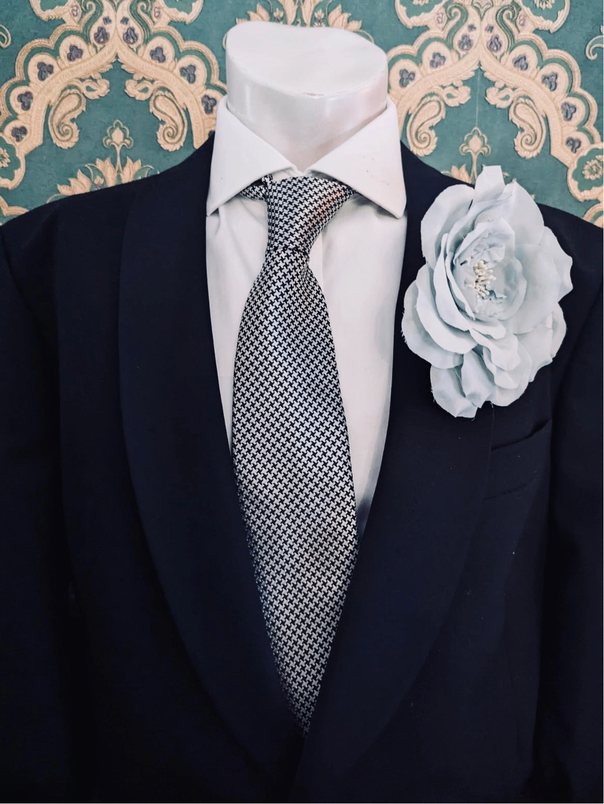 Broochella men's signorina fioritina handmade fabric flower brooch in sky blue. It is pinned on the label of a black suit jacket that is on a men's mannequin. The background is green and gold damask wallpaper.