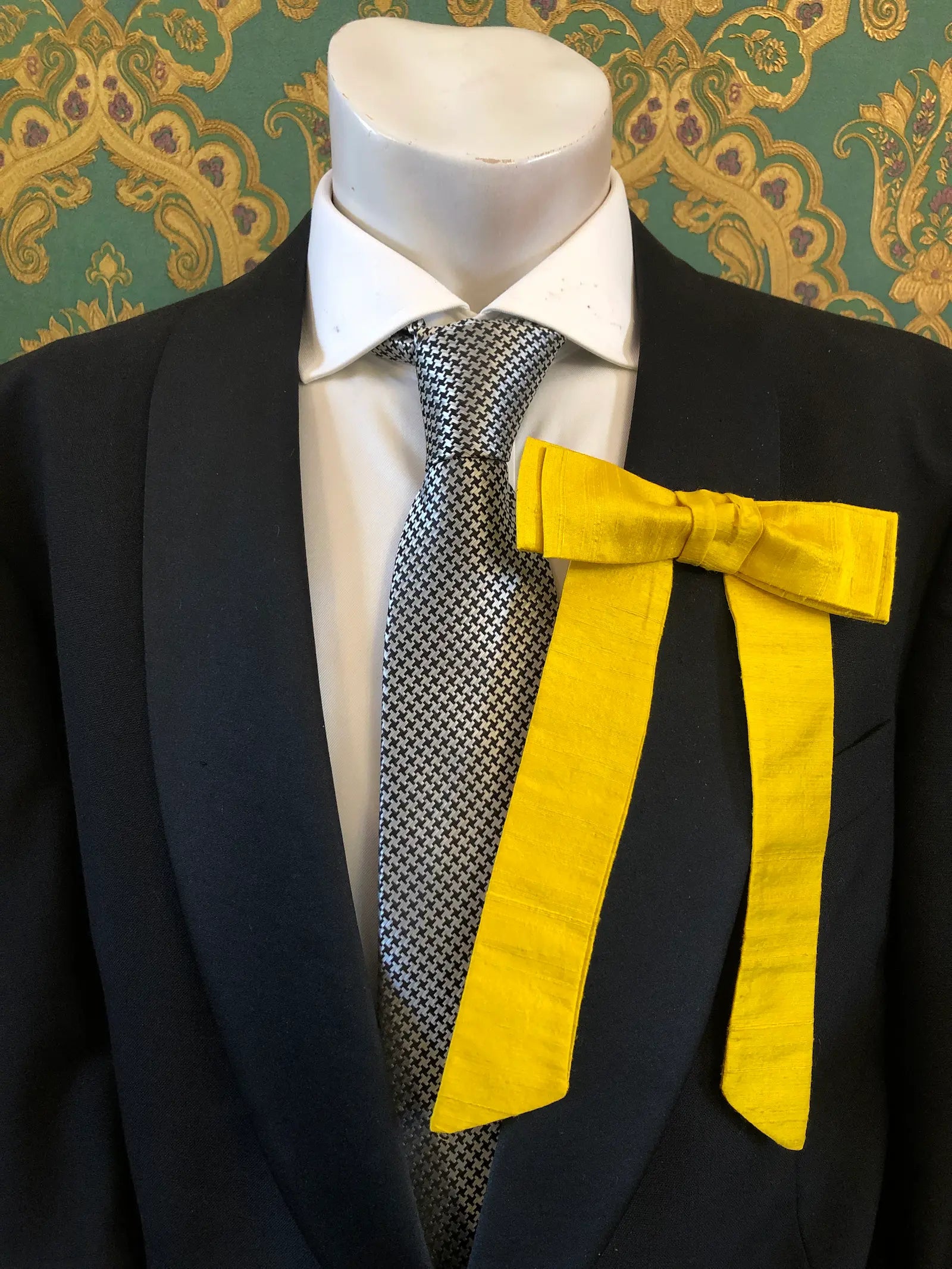 A Broochella Men's Alice silk bow brooch in yellow. It is styled on a white shirt and patterned tie, displayed on a black suit jacket mannequin torso against a green and gold damask wallpaper background.