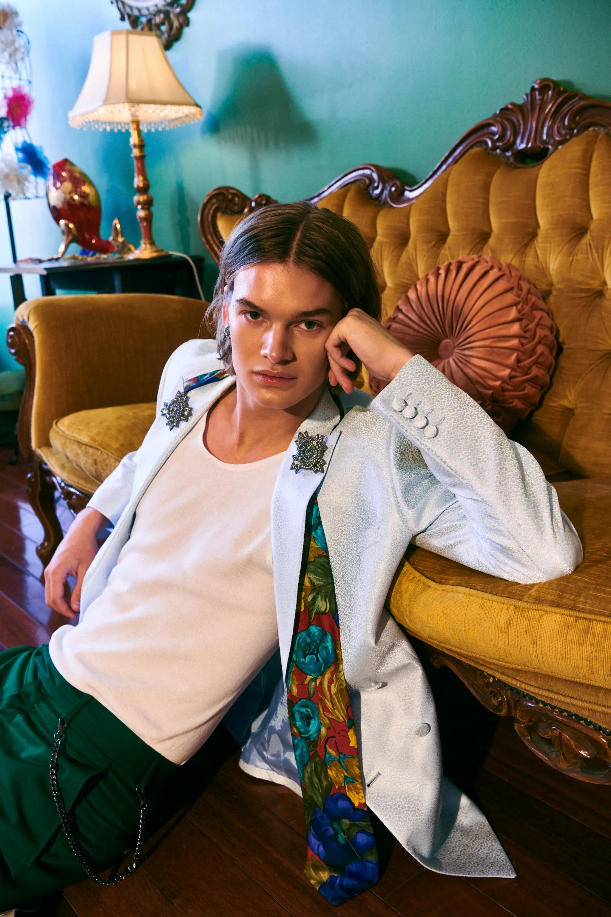 A model wearing a white blazer with Broochella men's silver starburst brooch lapel pins, a white t-shirt, green trousers, and a colorful floral necktie, leaning against a yellow tufted armchair in a vintage room.