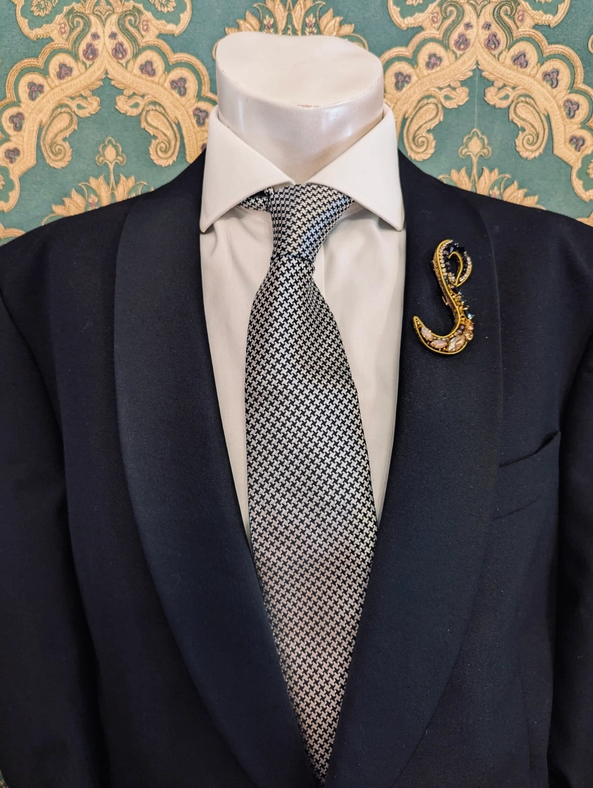 Broochella men's hand beaded S initial brooch in black and gold beads and sequins. Pinned to a men's mannequin wearing a black suit and a tie. The background is green and gold damask wallpaper.