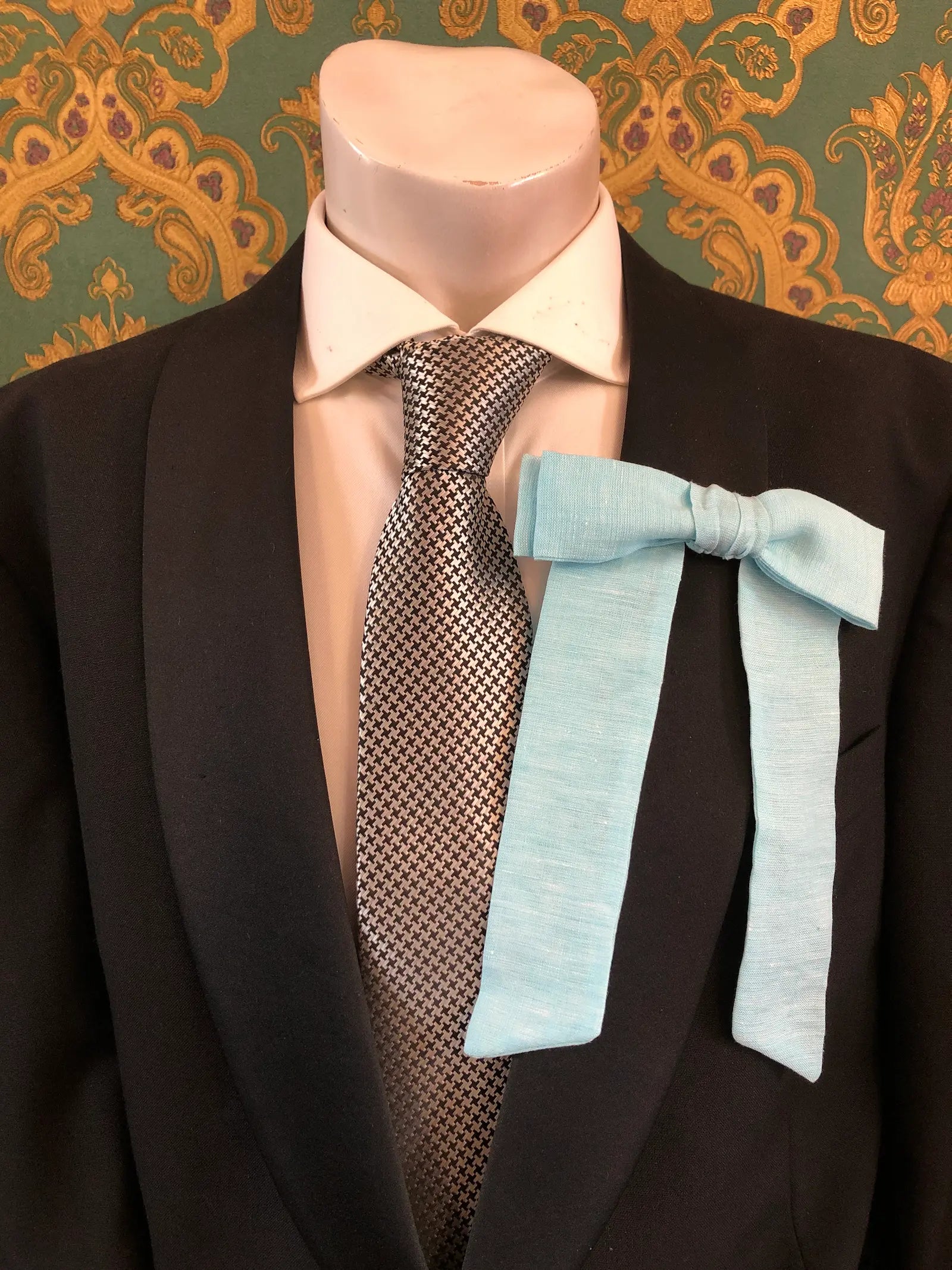 A Broochella Men's handmade Sofia Linen bow brooch in blue styled on a mannequin torso wearing a black suit jacket, white dress shirt, and a black-and-white checkered tie. The background is green and gold damask wallpaper. 