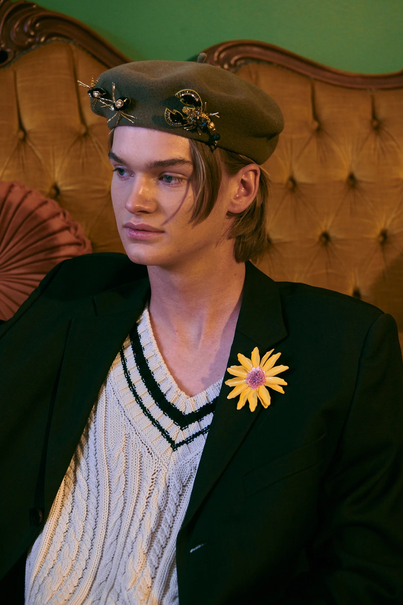 A model wearing a dark olive beret with gold star embellishments, a black blazer, and a cream knit sweater vest with a striped collar, seated on an ornate gold tufted armchair against a green wall. He is wearing the Broochella gold and black beaded spider brooch set on his beret.