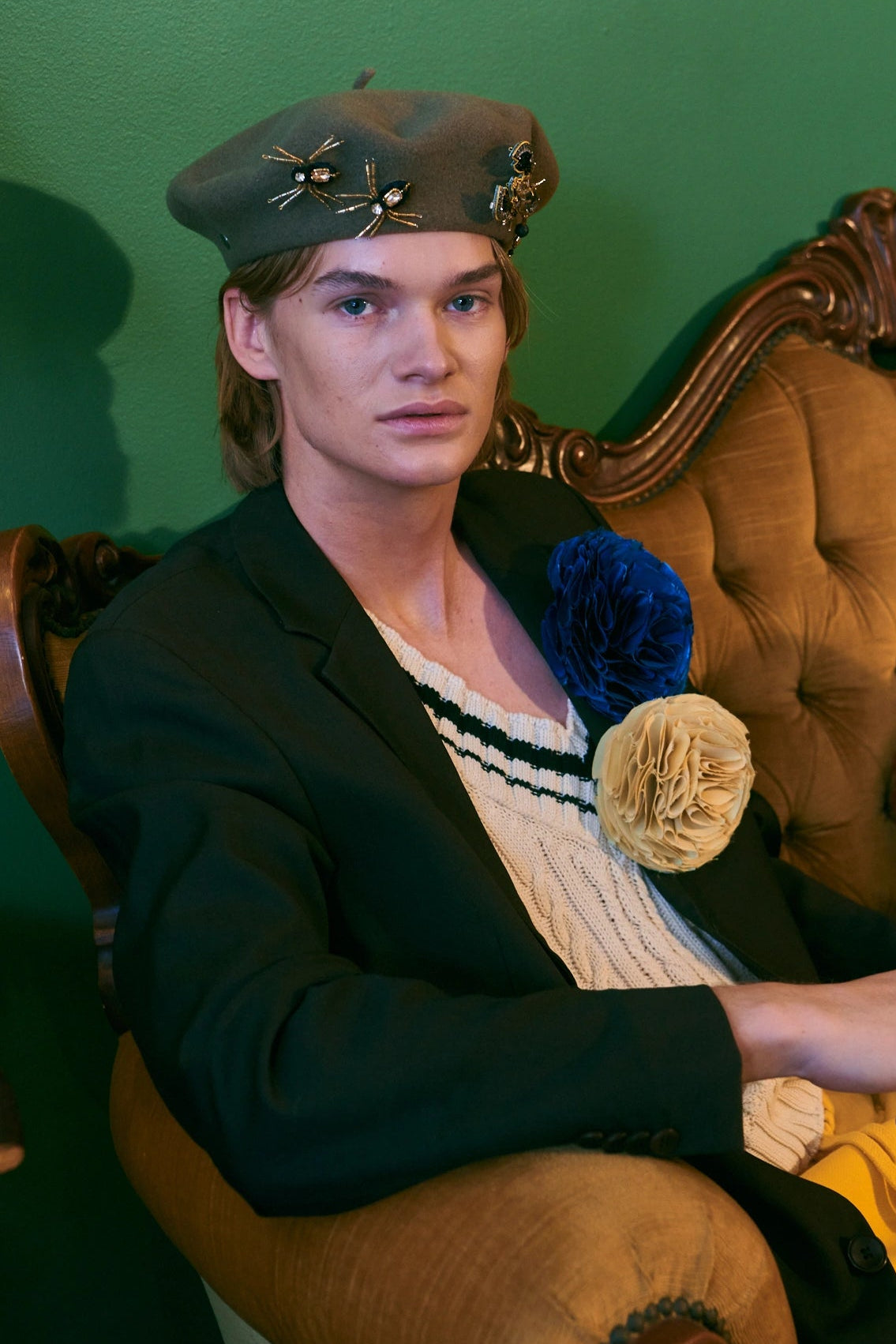 A model wearing a dark olive beret with gold star embellishments, a black blazer, and a cream knit sweater vest with a striped collar, seated on an ornate gold tufted armchair against a green wall. He is wearing the Broochella gold and black beaded spider brooch set on his beret.