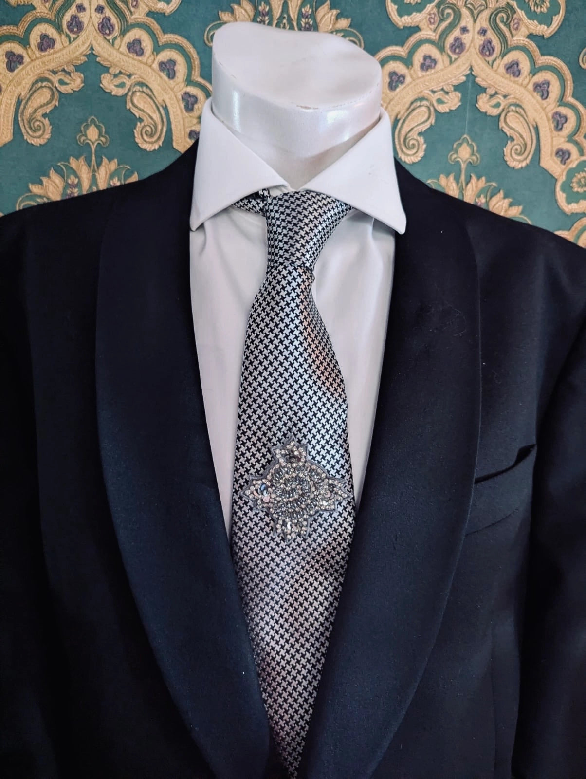 Broochella men's handmade, hand beaded designer, star bust brooch in beads and sequins. Pinned on a men's mannequin wearing a black suit and a houndstooth tie. The background is green and gold damask wallpaper. 