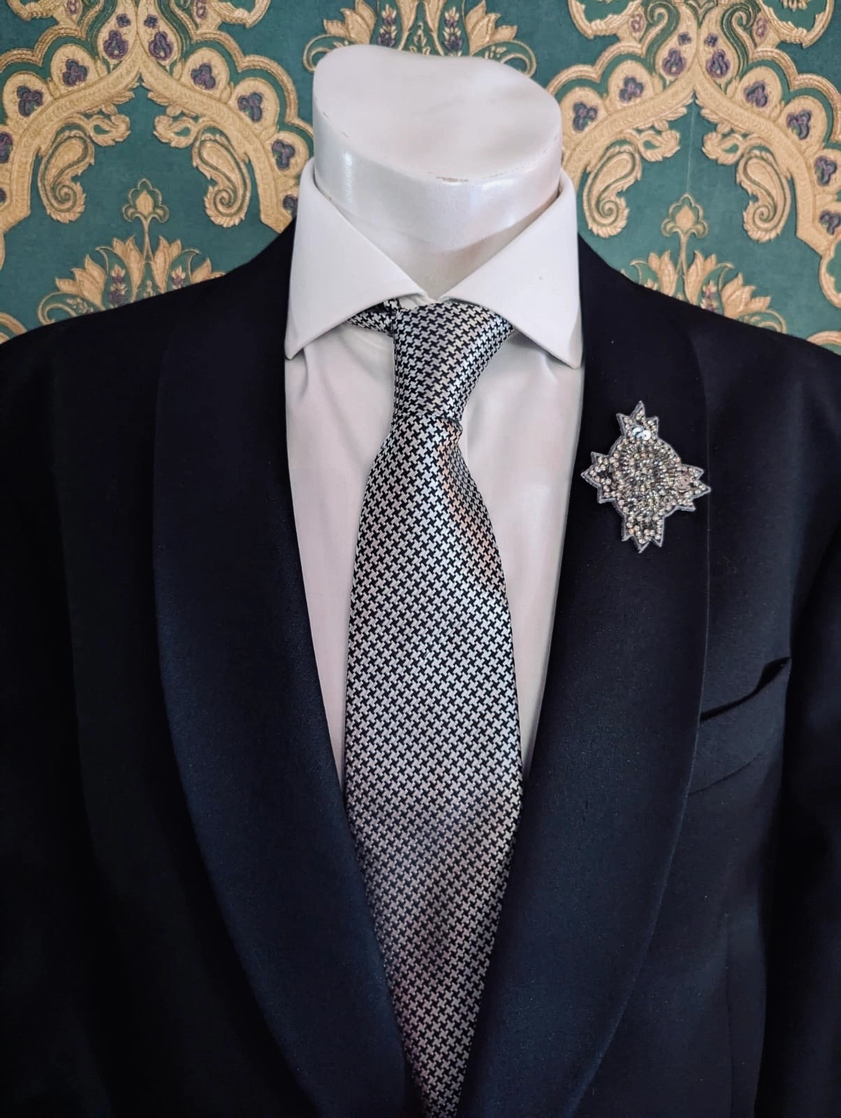 Broochella men's handmade, hand beaded designer, star bust brooch in beads and sequins. Pinned on a men's mannequin wearing a black suit and a houndstooth tie. The background is green and gold damask wallpaper. 