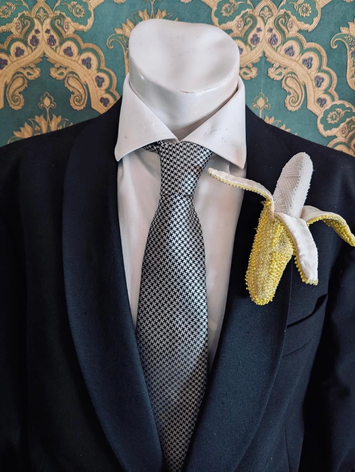 Mannequin wearing a suit with a white shirt, black tie, and a Broochella Tropicana handamde designer banana brooch against a decorative wall.