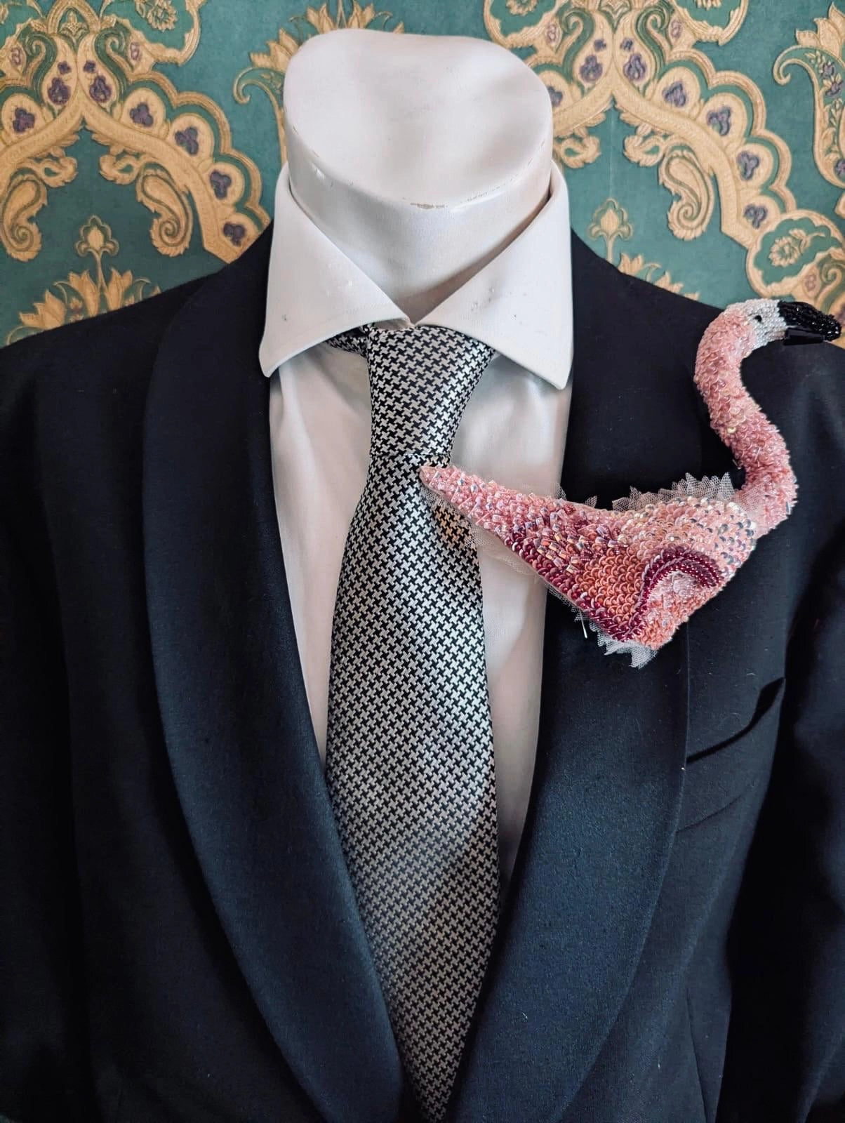 A Broochella men's Tropicana pink beaded flamingo brooch pinned to the lapel of a black suit jacket, styled with a white dress shirt and a grey patterned tie on a mannequin torso. The background is green and gold damask wallpaper.