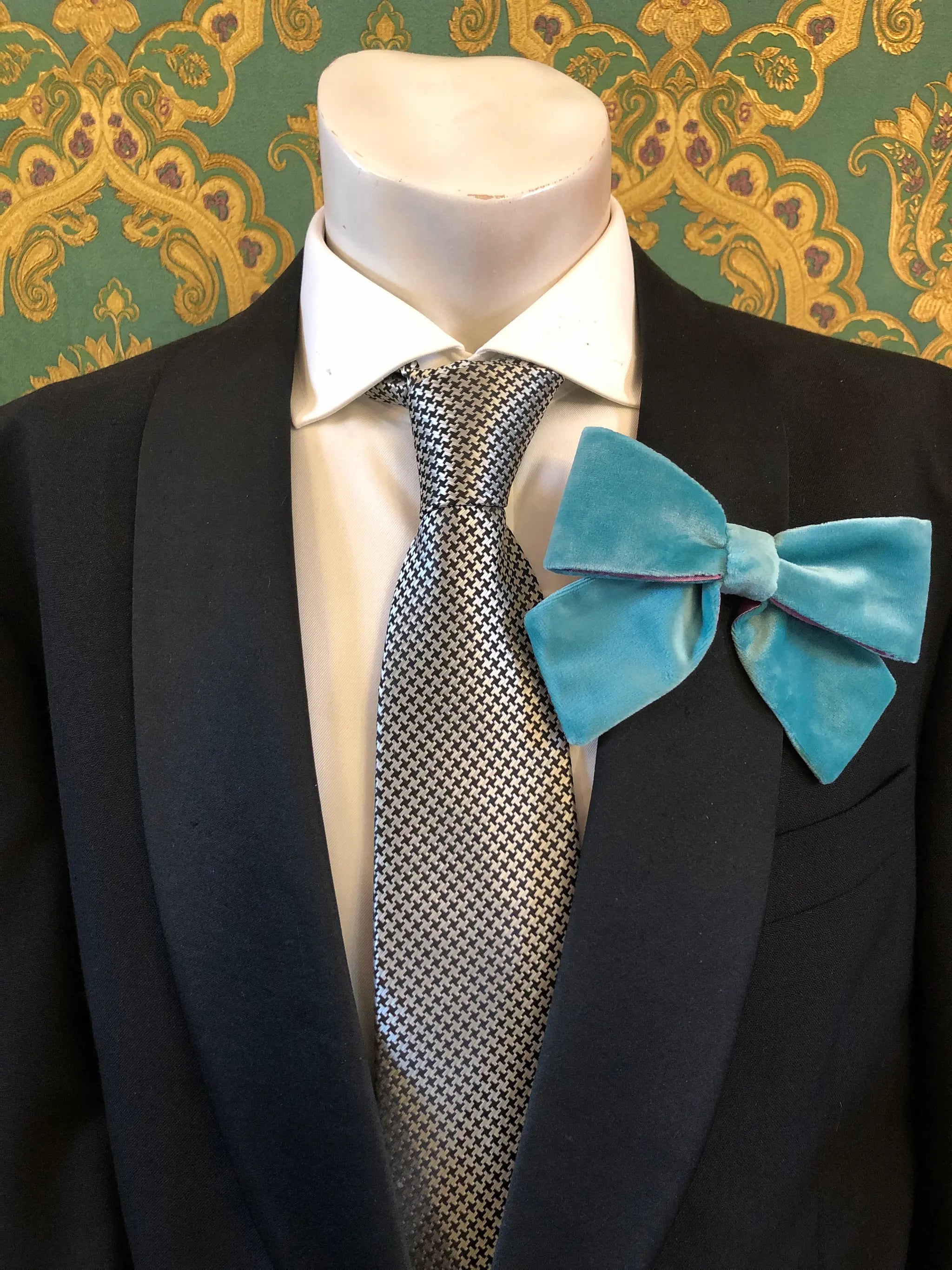 A Broochella Men's aqua/purple velvet bow brooch displayed on a mannequin torso wearing a black suit jacket, white dress shirt, and a silver-and-black patterned tie. It is set against ornate green and gold patterned wallpaper.