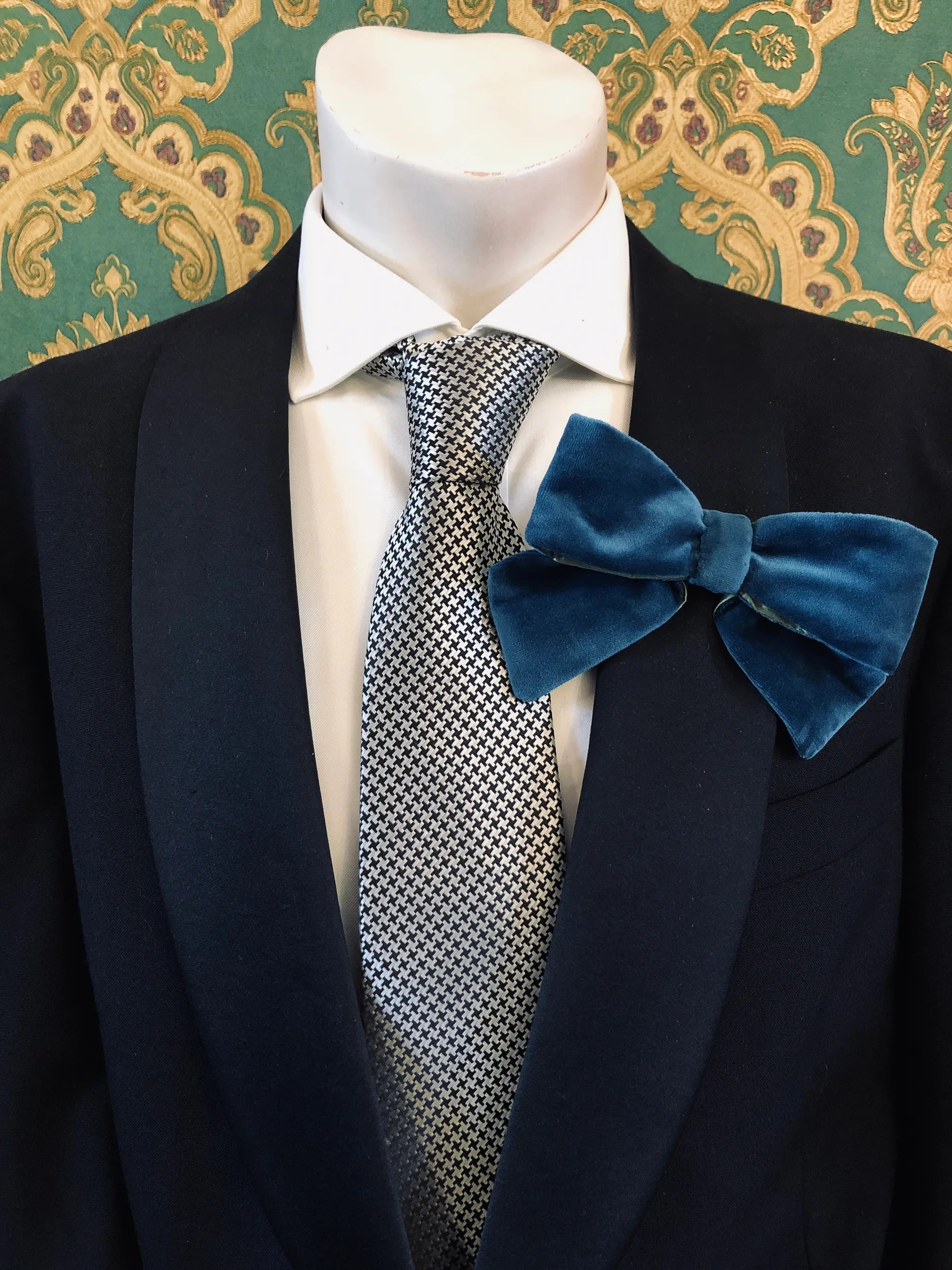 A Broochella Men's peacock blue/green velvet bow brooch displayed on a mannequin torso wearing a black suit jacket, white dress shirt, and a silver-and-black patterned tie. It is set against ornate green and gold patterned wallpaper.