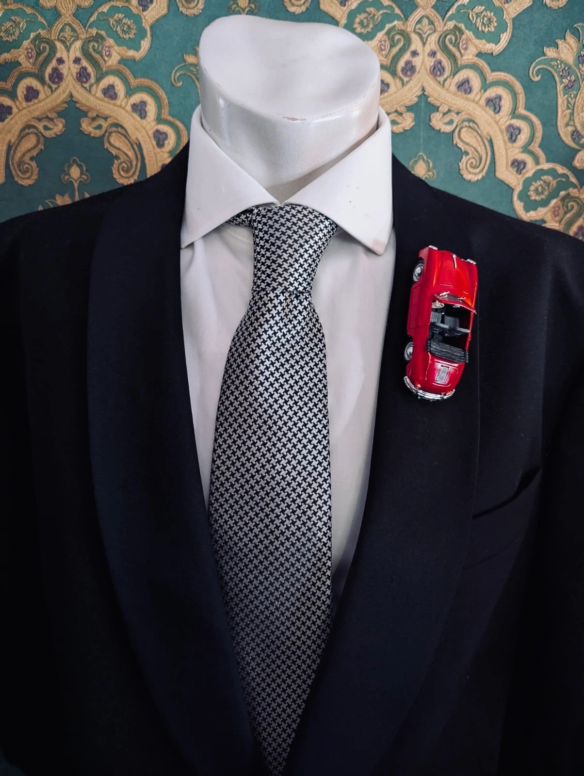 Broochella men's scuderia d'oro rosso cabrio number 356 red, vintage formula one racing car brooch. Pinned on a men's mannequin wearing a black suit and a houndstooth tie. The background is green and gold damask wallpaper. 