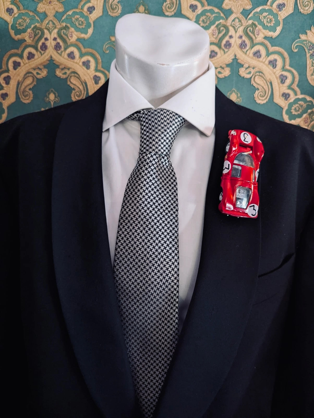 Broochella men's scuderia d'oro rosso le mans number 3 vintage formula one racing car brooch. Pinned on a men's mannequin wearing a black suit and a houndstooth tie. The background is green and gold damask wallpaper. 