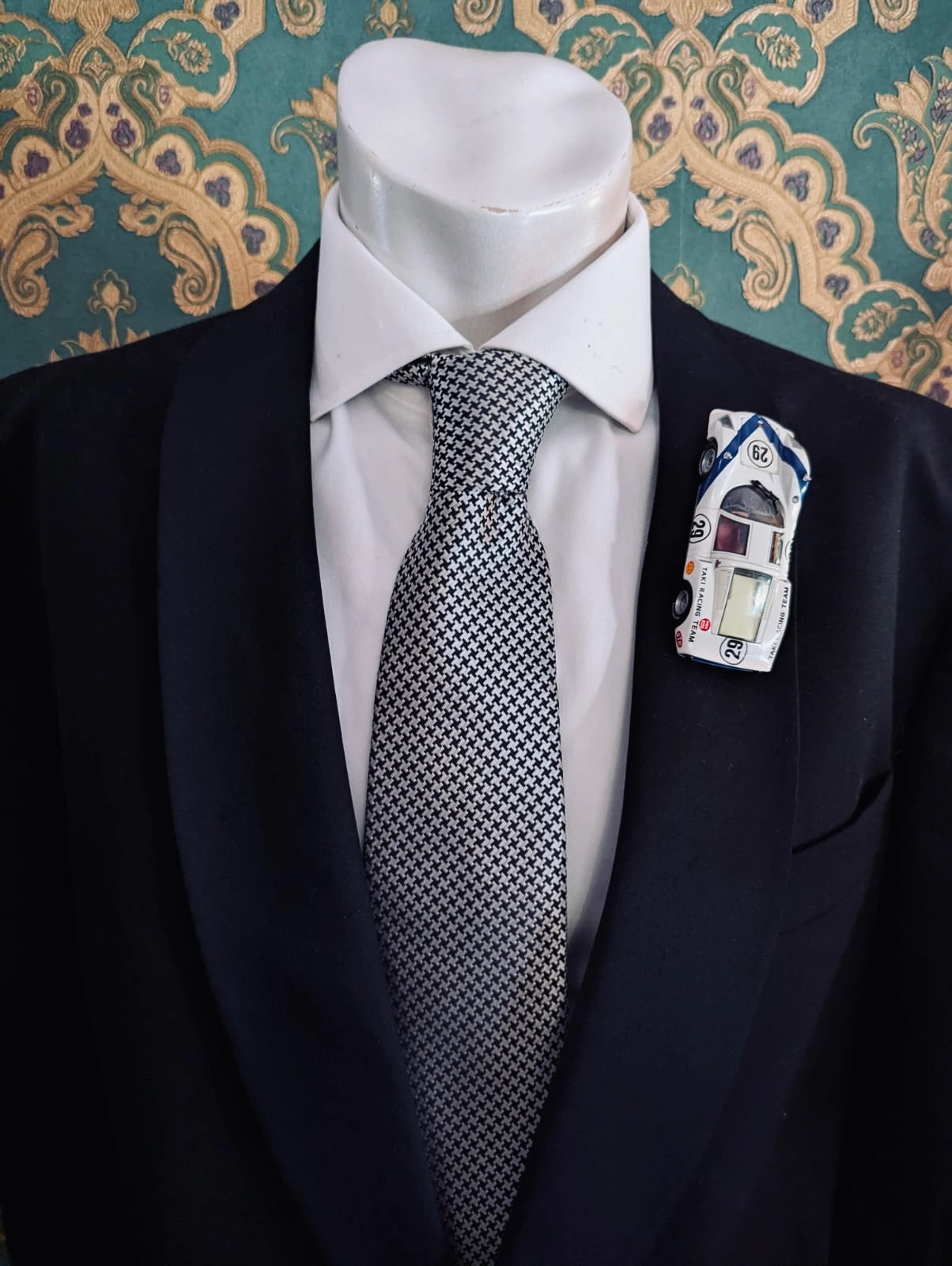 Broochella men's scuderia d'oro bianco takumi number 29 vintage formula one racing car brooch. Pinned on a men's mannequin wearing a black suit and a houndstooth tie. The background is green and gold damask wallpaper. 
