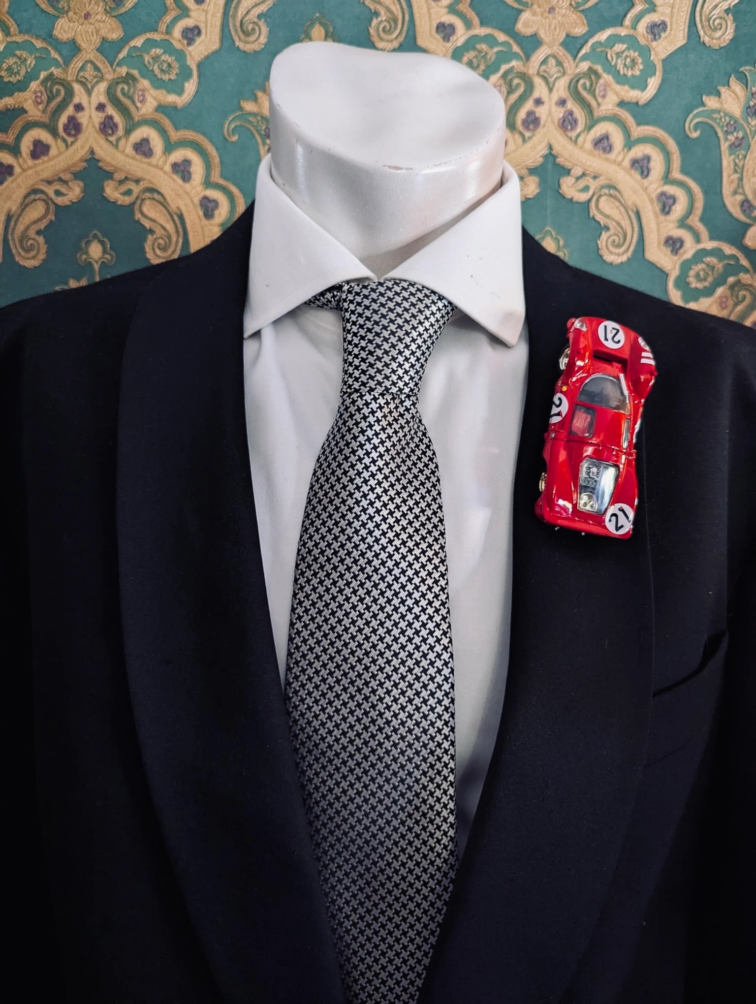 Broochella men's scuderia d'oro rosso fiorentino number 21 red, vintage formula one racing car brooch. Pinned on a men's mannequin wearing a black suit and a houndstooth tie. The background is green and gold damask wallpaper. 