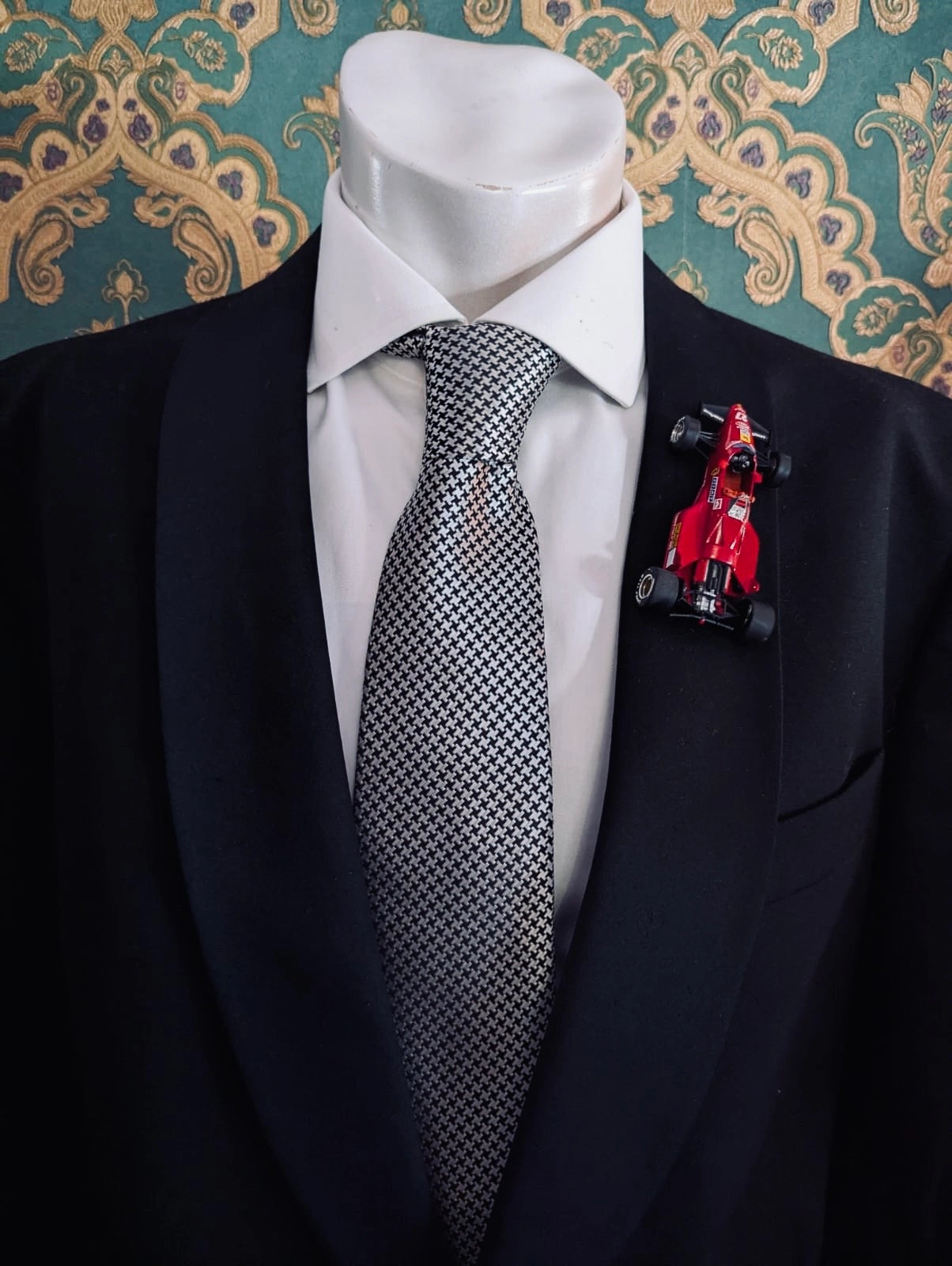 Broochella men's scuderia d'oro rosso maranello number 27 red,  vintage formula one racing car brooch. Pinned on a men's mannequin wearing a black suit and a houndstooth tie. The background is green and gold damask wallpaper. 