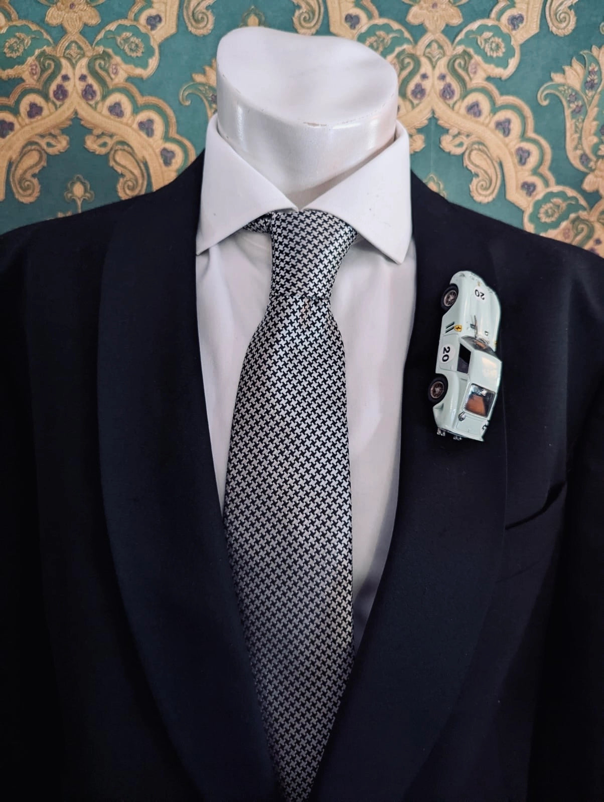 Broochella men's scuderia d'oro verde aqua number 20 vintage formula one racing car brooch. Pinned on a men's mannequin wearing a black suit and a houndstooth tie. The background is green and gold damask wallpaper. 
