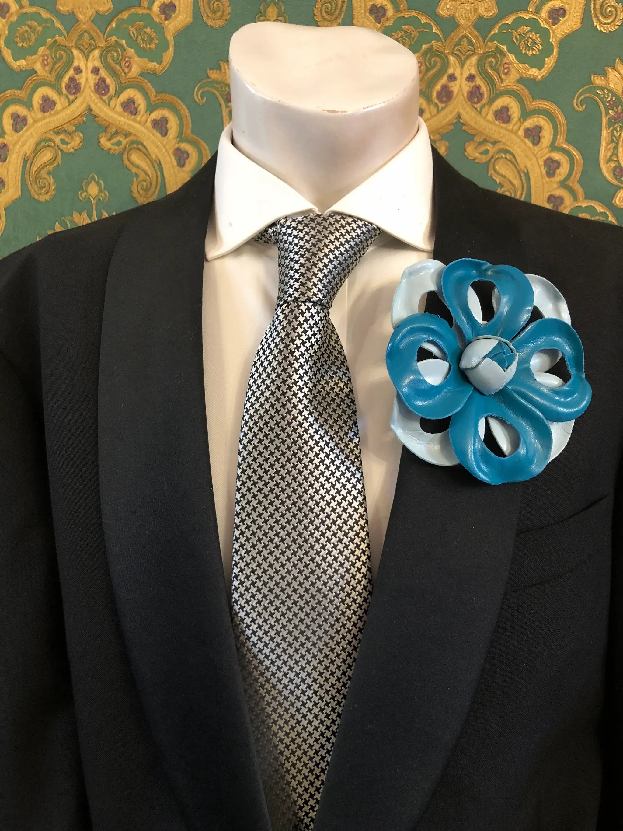 A Broochella Men's Viola del Pensiero handmade designer blue leather flower-shaped brooch pin. It is displayed  on a black suit jacket, white dress shirt, and patterned grey tie on a mannequin torso The background is ornate green and gold patterned wallpaper.