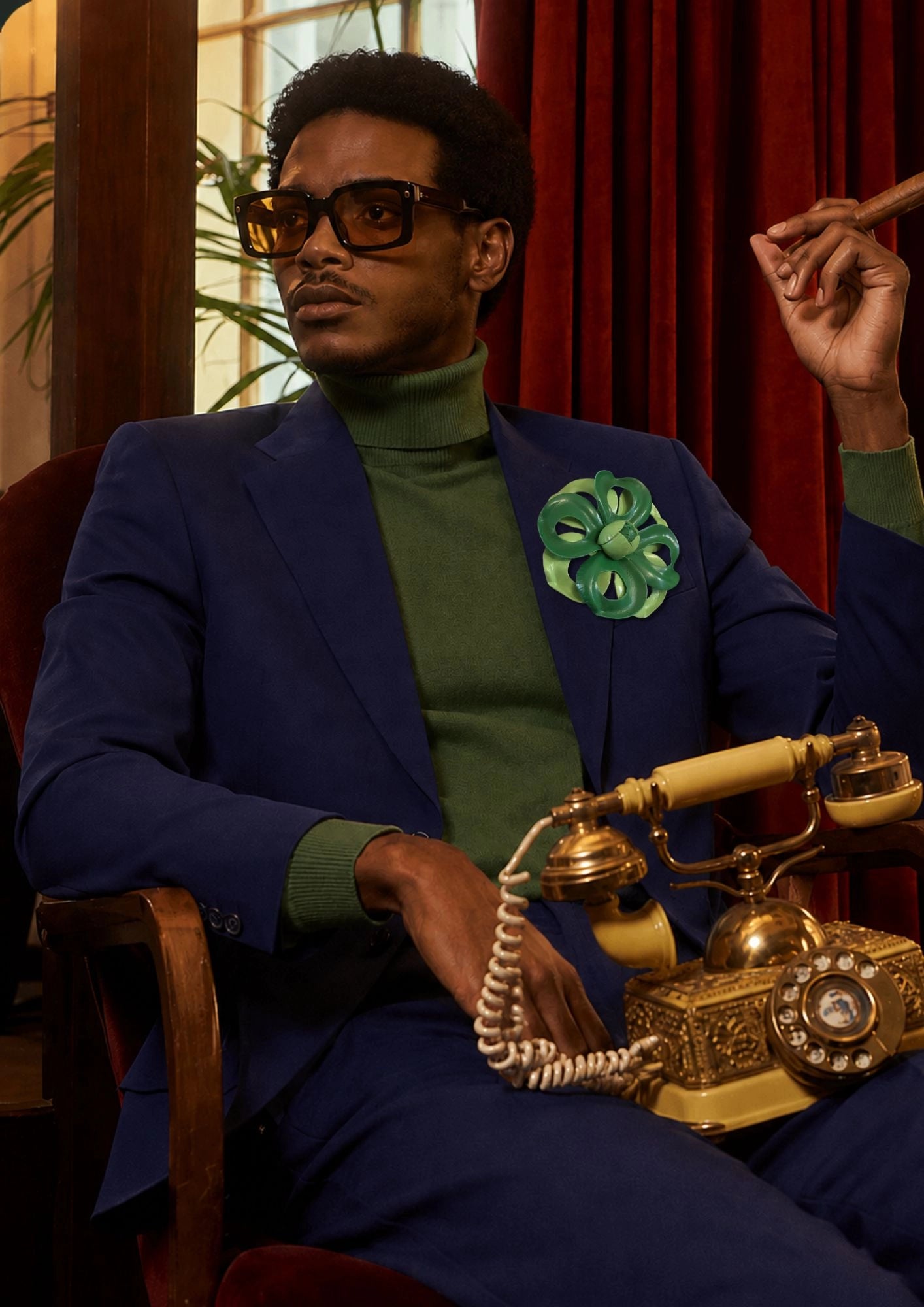 A dark haired man in a blue suit and green skivvy is sitting with an old phone in his lap and a cigar in his hand. The background is red velvet curtains. Pinned to his lapel is the Broochella men's viola del pensiero leather flower brooch in green. 