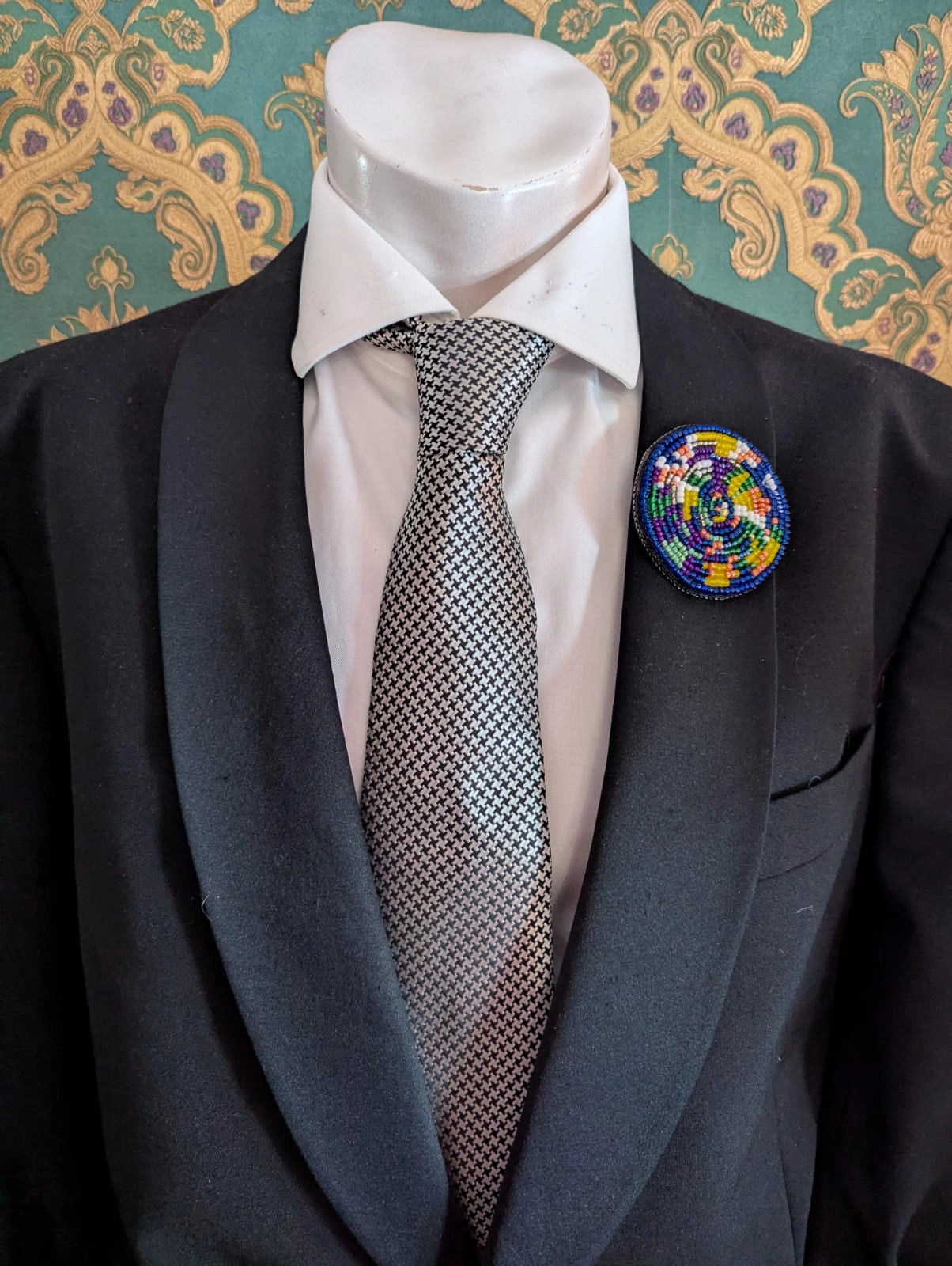 Broochella  men's viva la gioia hand beaded brooch. The brooch is round, with spirals of multicoloured beads. The brooch is pinned on a men's mannequin in a black suit The background is green and gold damask wallpaper.