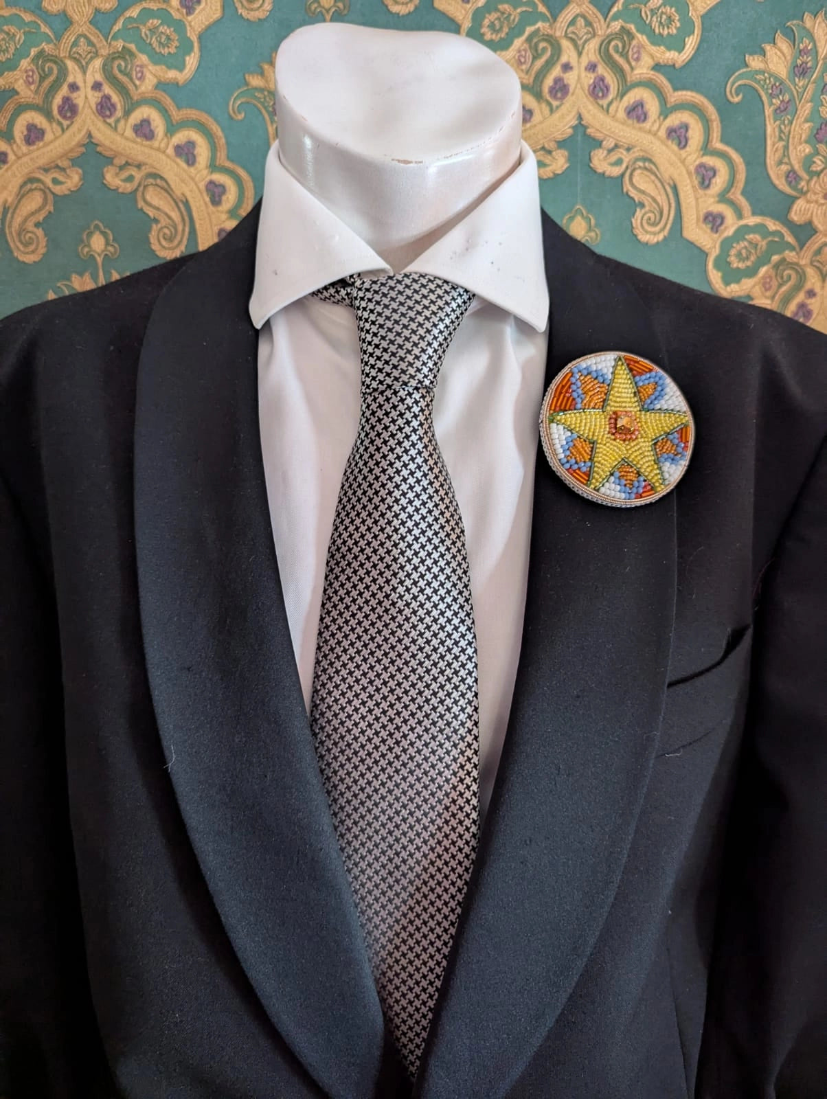 Broochella  men's viva la stella hand beaded brooch. The brooch is round, with a yellow star at the centre. There are blue, orange and white beads around the star. The brooch is pinned on a men's mannequin in a black suit The background is green and gold damask wallpaper.
