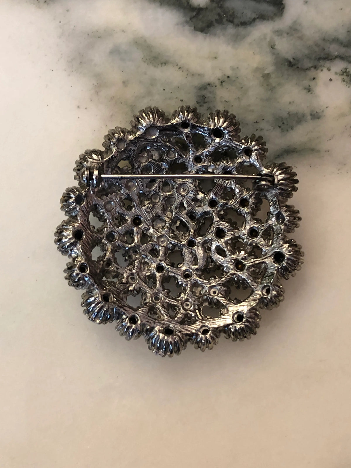 The back view Broochella Men's Alessia Vintage Black & Crystal Brooch featuring clear crystals and jet-black stones in an ornate silver-tone circular setting on a cream and grey marble background.