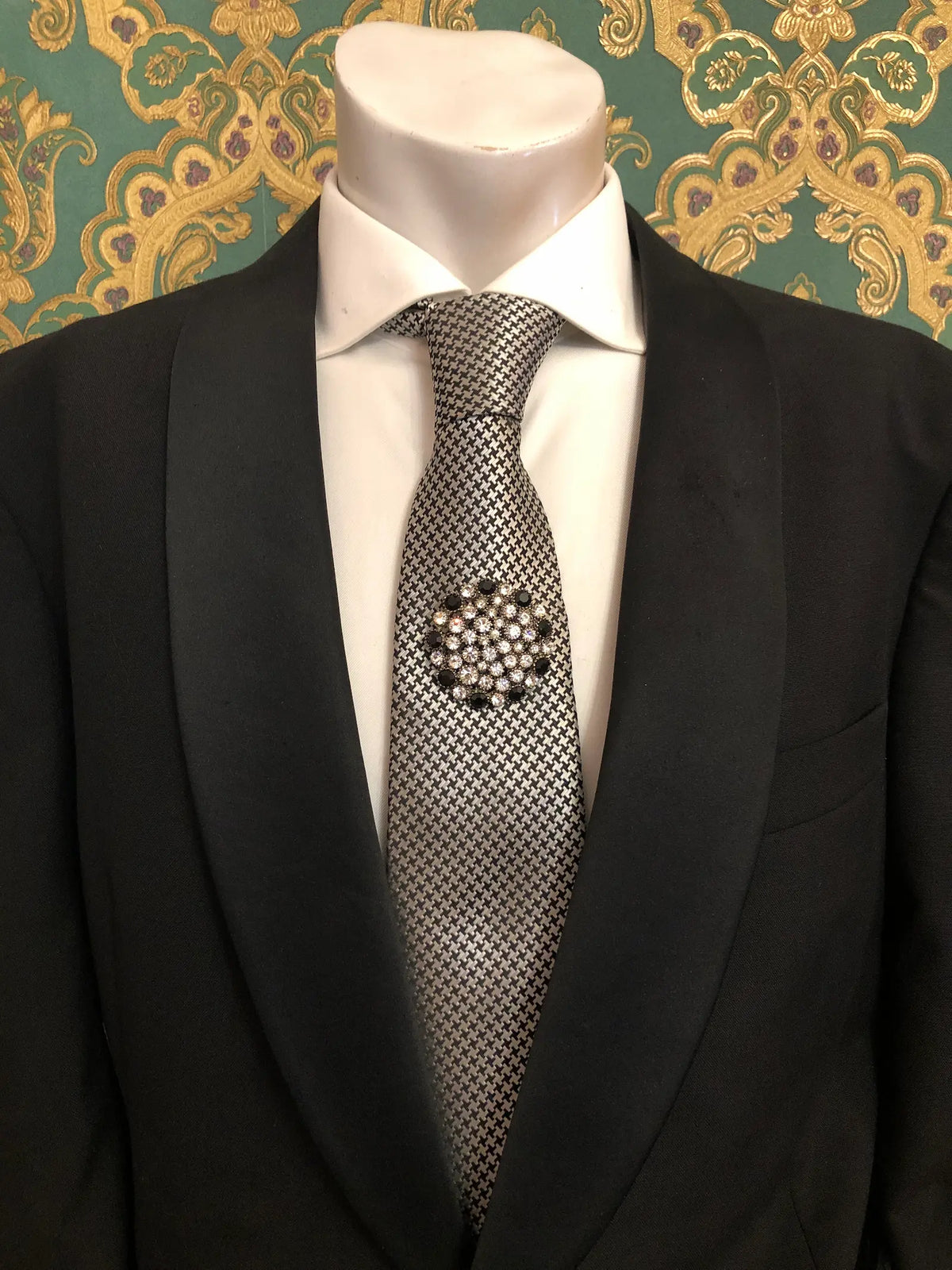 Broochella Men's Alessia vintage black and crystal brooch featuring a round arrangement of shimmering clear rhinestones accented with black stones, styled on a black-and-white patterned necktie with a crisp white shirt and black tailored suit, photographed against decorative emerald green and gold damask wallpaper.