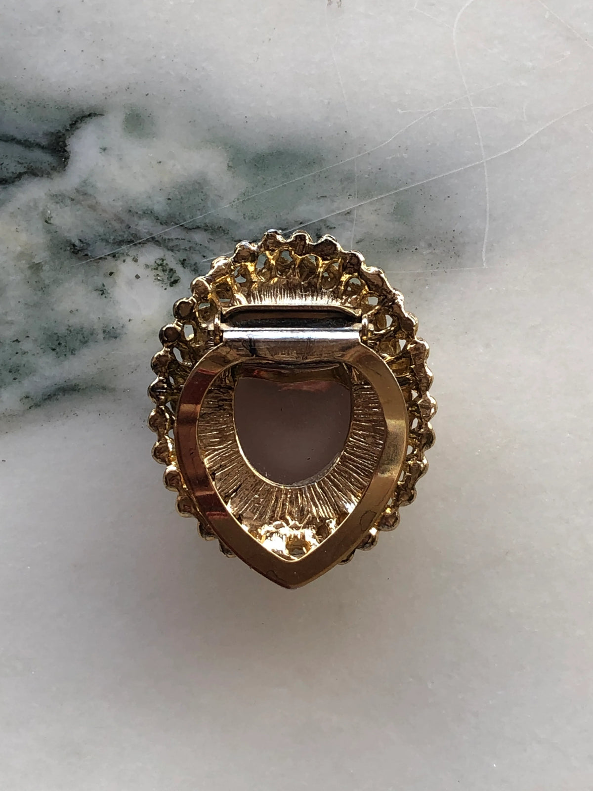 Broochella Men’s Amber Crest Vintage Tie Clip featuring a polished amber-brown oval cabochon framed by an ornate textured gold-tone setting, photographed on white marble with natural grey-green veining.