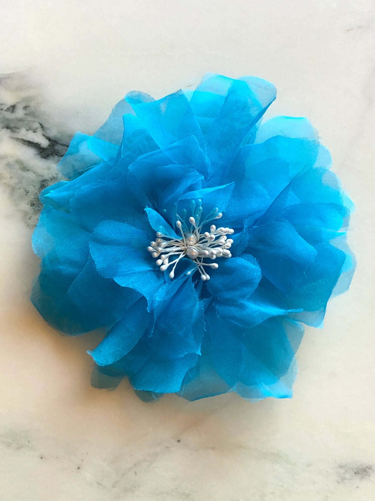 Broochella men’s Aubrieta handmade designer flower brooch in vibrant blue fabric with pearl stamen detailing, photographed on a white and green veined marble background.