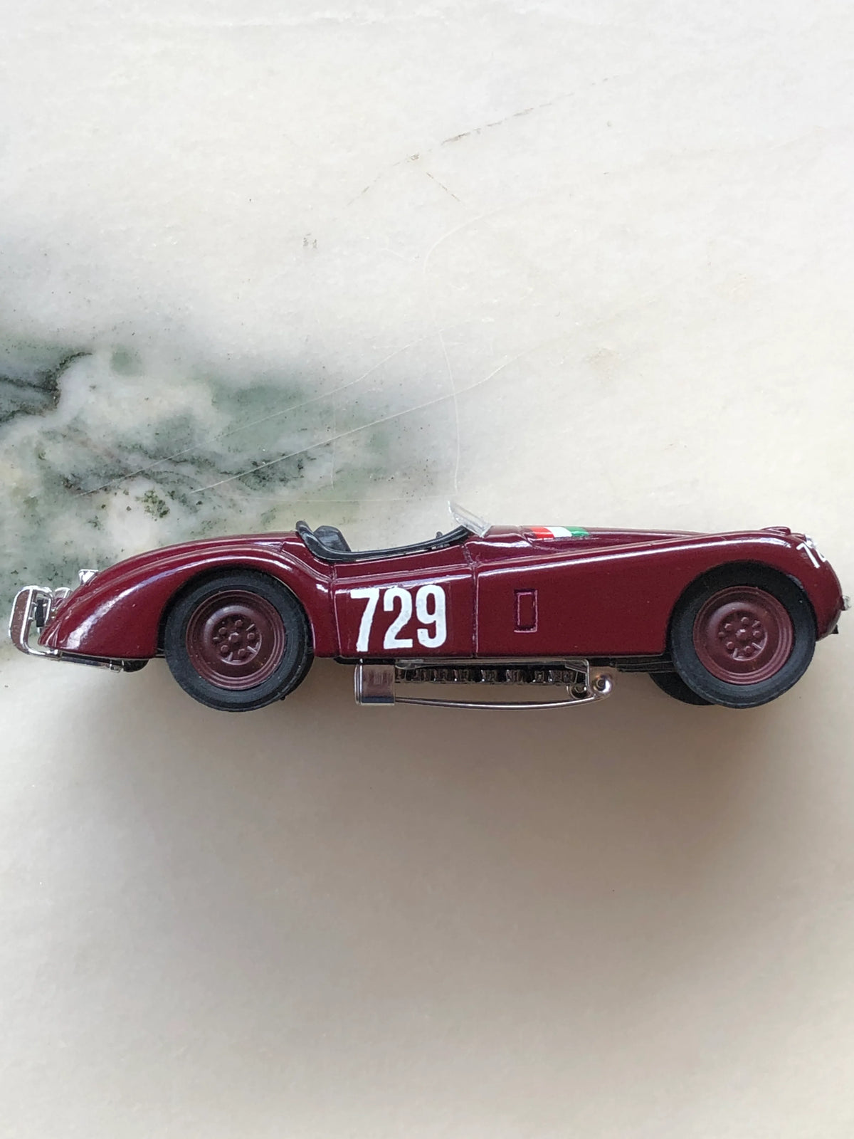 Broochella Men's Barolo No.729 Vintage Racing Car Brooch in side profile, featuring a streamlined burgundy roadster body, white number 729 racing decal, matching burgundy wheels, open cockpit and visible metal brooch pin fastening.