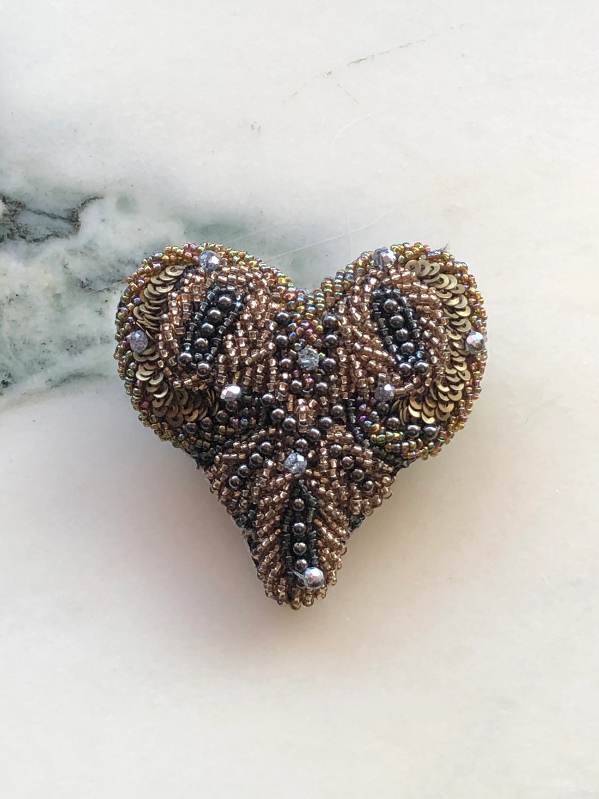 Broochella Men’s Beaded Amore Heart Brooch featuring an ornate heart design hand embellished with bronze, gold and gunmetal beads, sequins and sparkling crystals, photographed on a white marble background with grey-green veining.