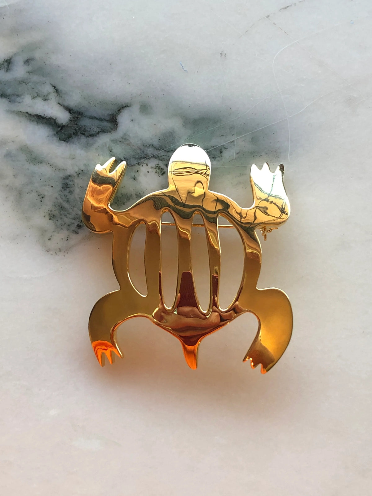 Broochella Men’s Brooches with Meaning Adaptability Brooch featuring an abstract polished gold-tone frog design, photographed on a pale white marble background with grey veining.