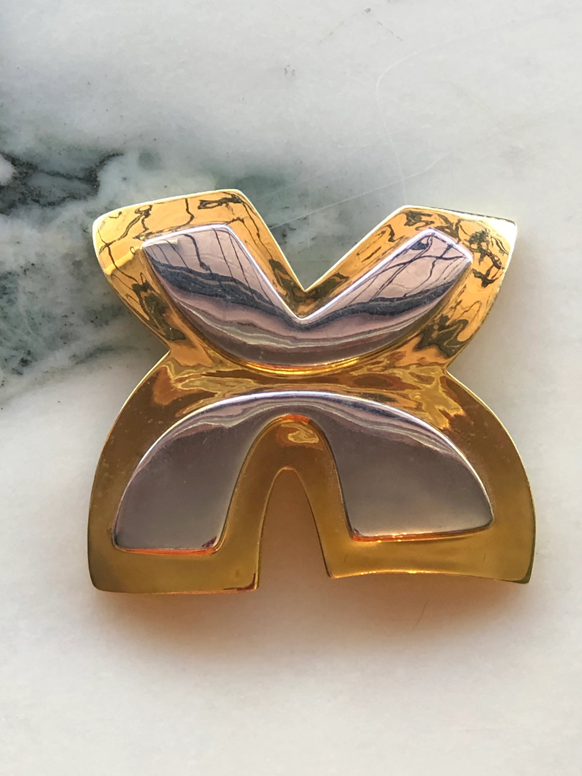Broochella Men’s Brooches with Meaning Freedom Brooch featuring an abstract sculptural design in polished gold and silver tones, photographed on a pale white marble background with grey veining.