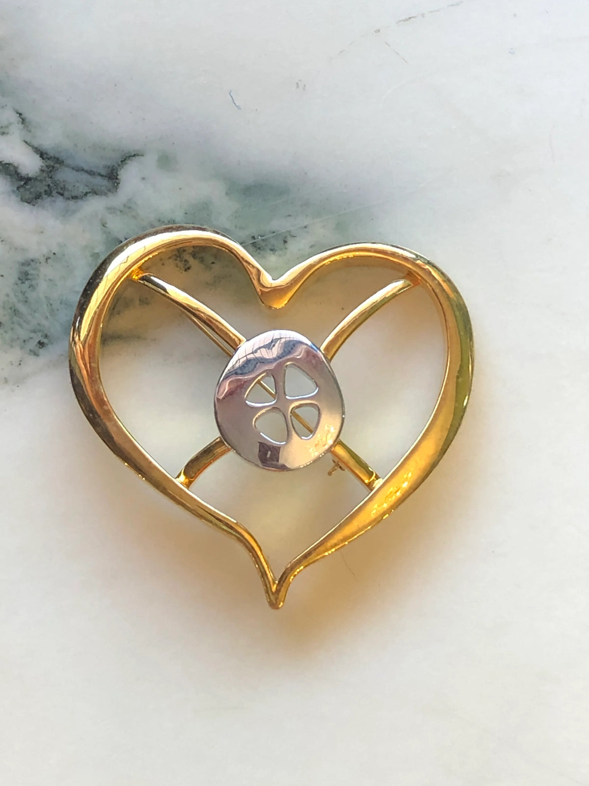 Broochella Men’s Brooches with Meaning Hope Brooch featuring an open heart design in polished gold with a sculptural silver-tone centre, photographed on a pale white marble background with grey veining.