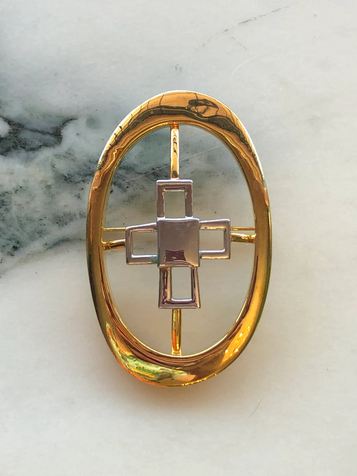 Broochella Men’s Brooches with Meaning Knowledge Brooch featuring an open polished gold oval frame with a geometric silver-tone cross motif at the centre, photographed on a pale white marble background with grey veining.