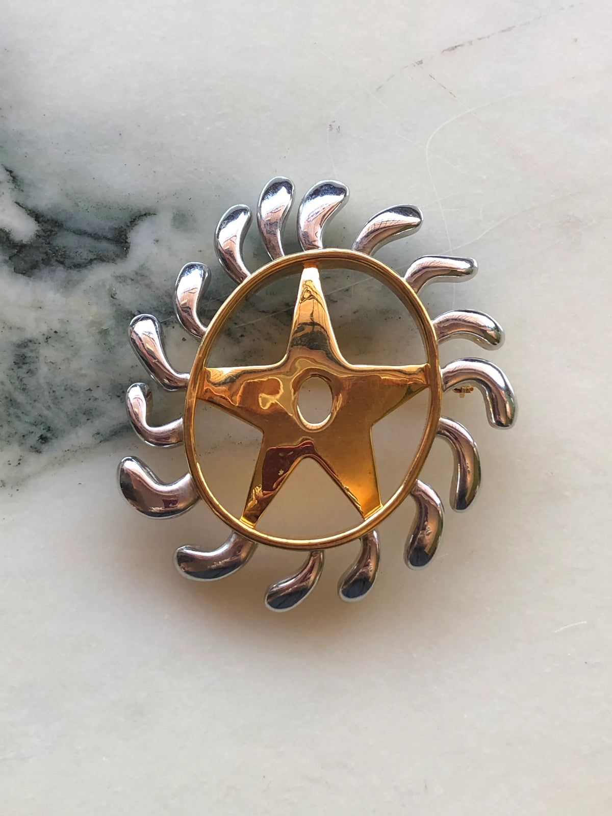 Broochella Men’s Brooches with Meaning Welcoming Change Brooch featuring a polished gold human figure within a circular frame surrounded by sculptural silver-tone rays, photographed on a pale white marble background with grey veining.