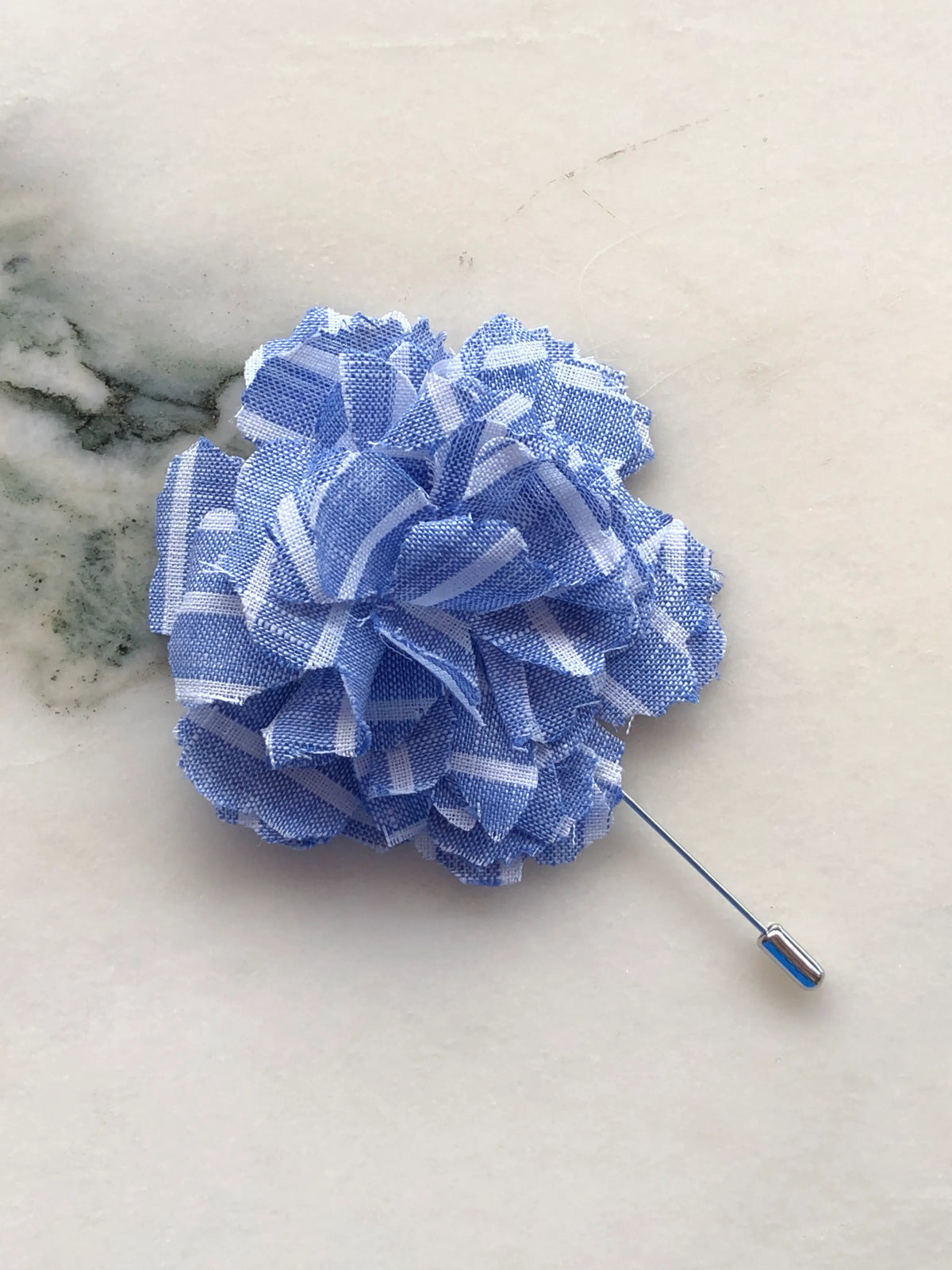 Broochella Men’s Carnation Flower Brooch in blue patterned fabric with layered petals and silver stick pin, photographed on a white marble background with grey-green veining.