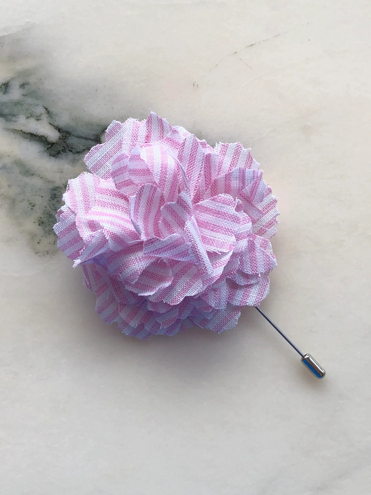 Broochella Men’s Carnation Flower Brooch in blush pink striped fabric with layered petals and silver stick pin, photographed on a white marble background with grey-green veining.