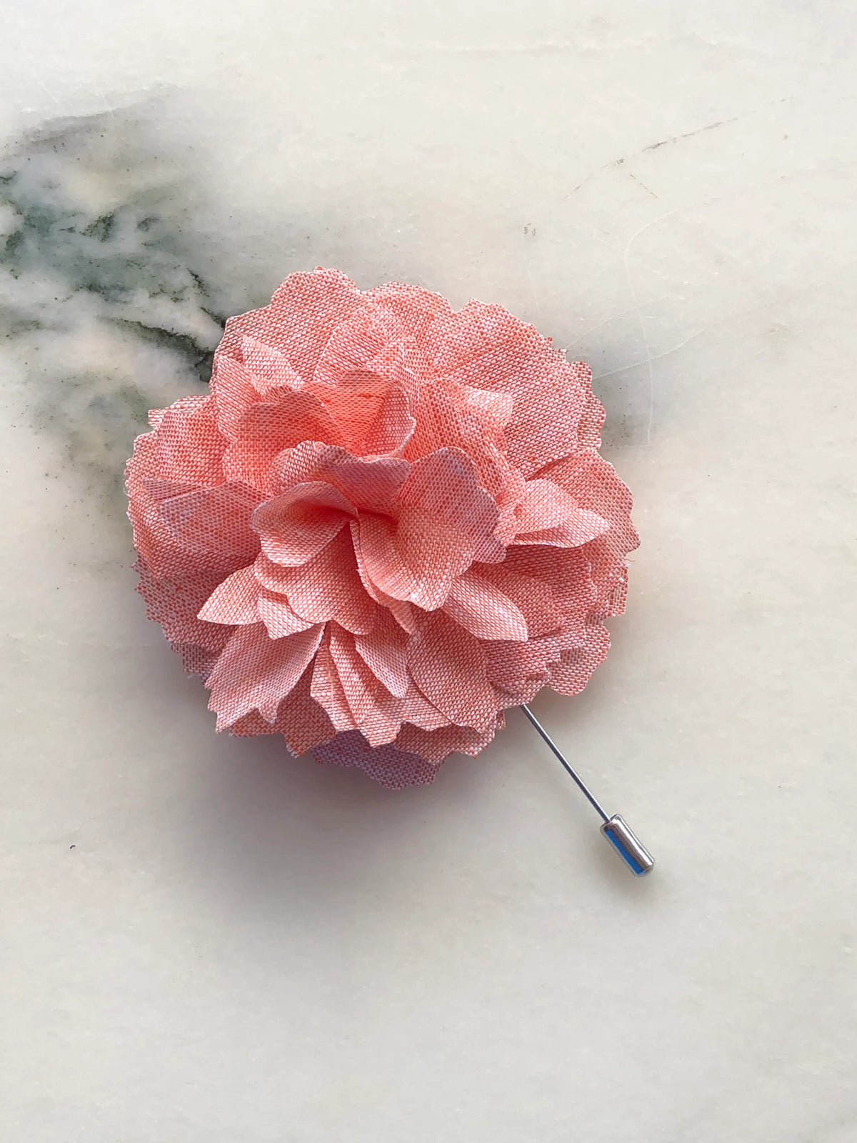 Broochella Men’s Carnation Flower Brooch in soft orange fabric with layered petals and silver stick pin, photographed on a white marble background with grey-green veining.
