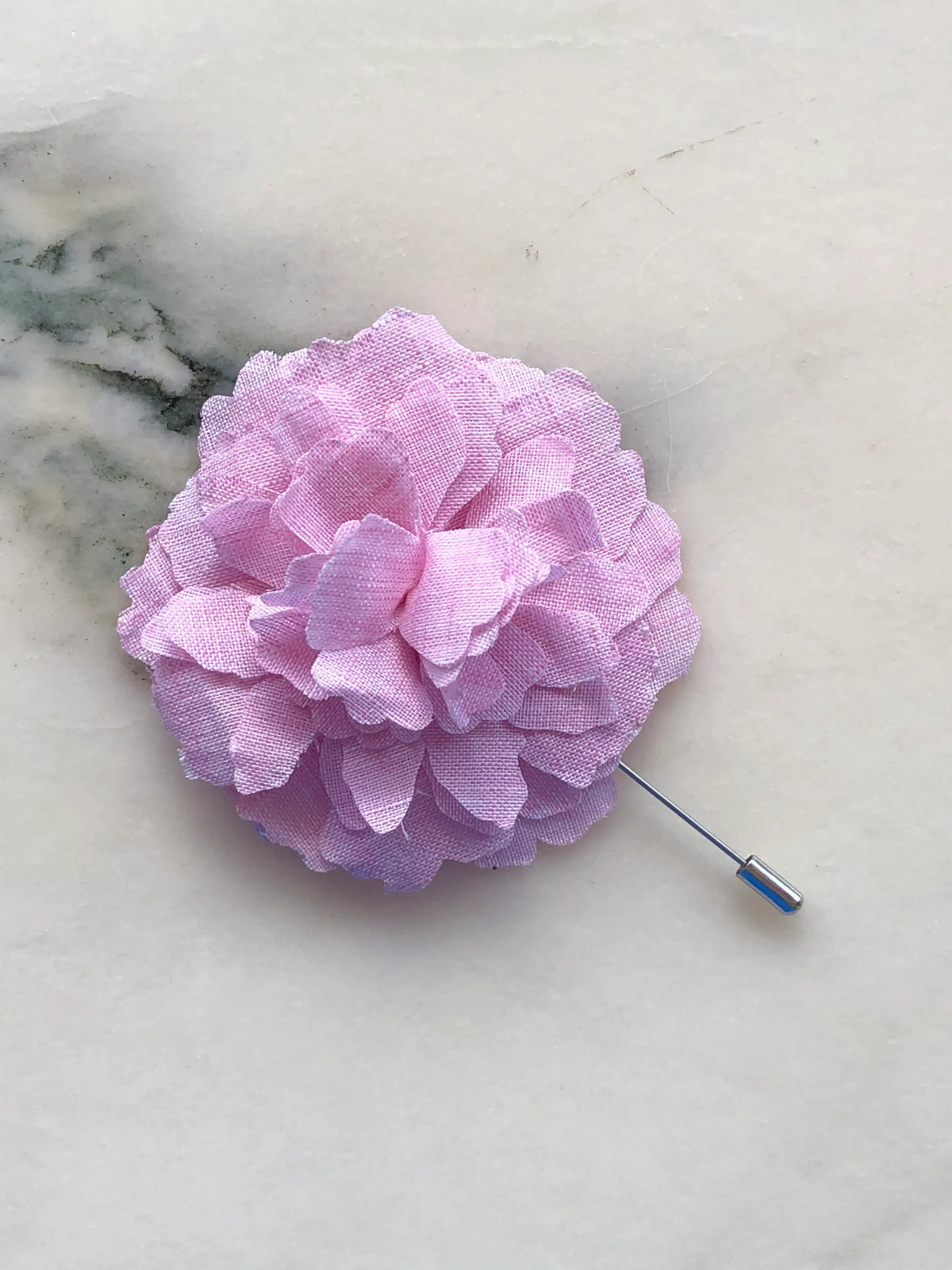 Broochella Men’s Carnation Flower Brooch in soft pink fabric with layered petals and silver stick pin, photographed on a white marble background with grey-green veining.