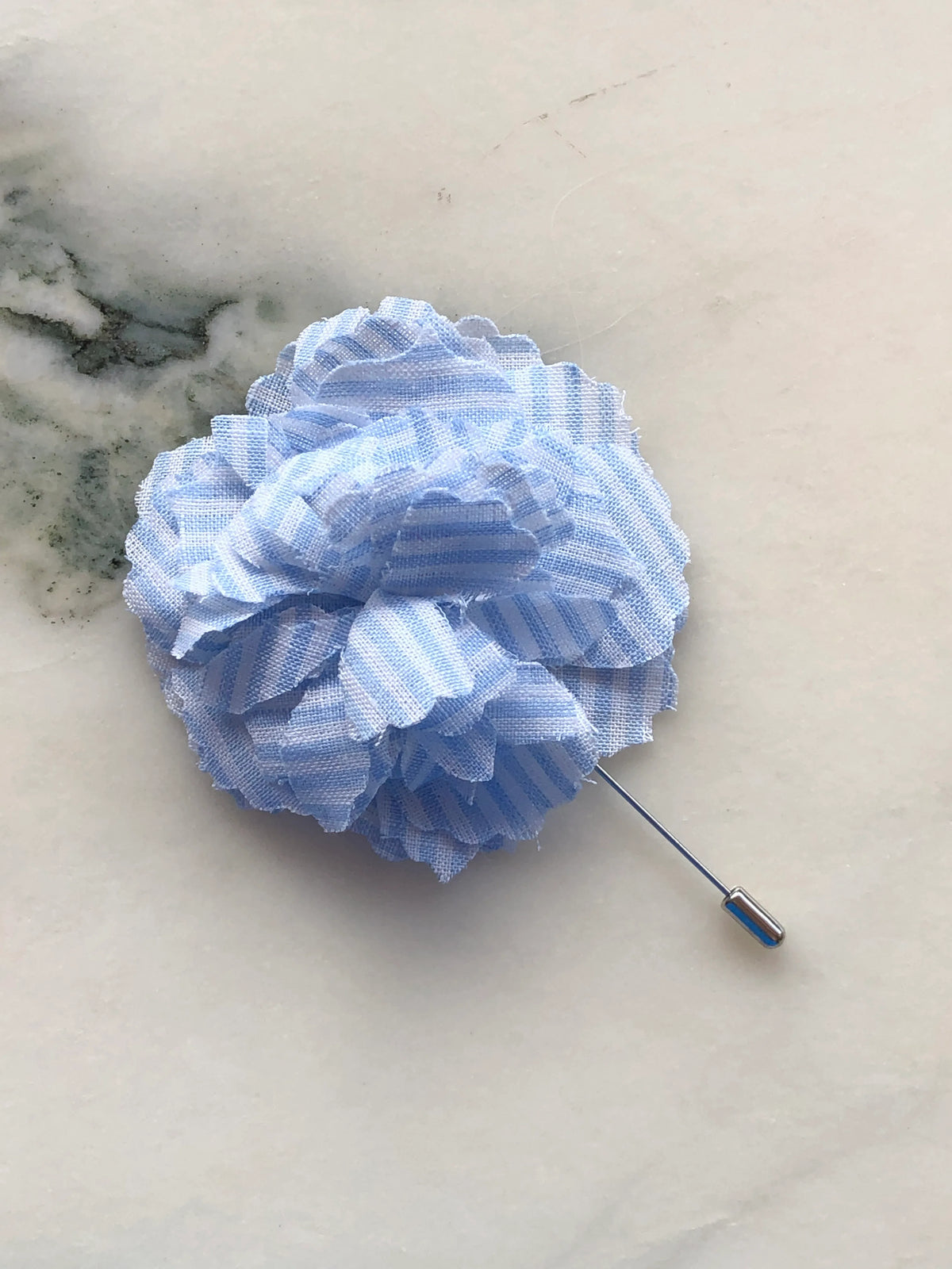 Broochella Men’s Carnation Flower Brooch in sky blue striped fabric with layered petals and silver stick pin, photographed on a white marble background with grey-green veining.