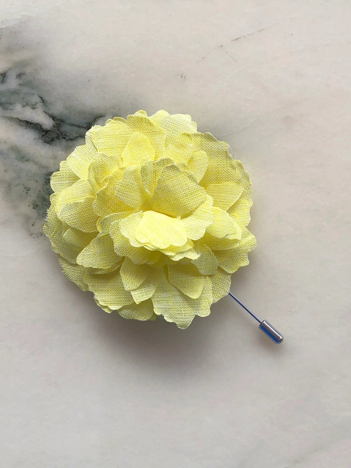 Broochella Men’s Carnation Flower Brooch in fresh yellow fabric with layered petals and silver stick pin, photographed on a white marble background with grey-green veining.