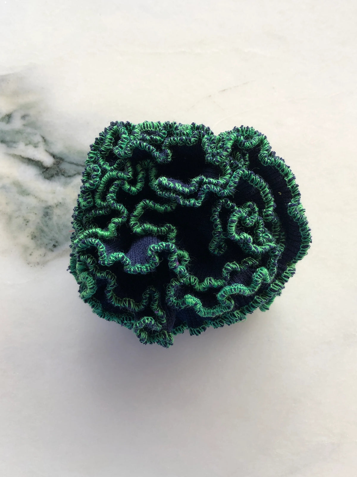 Broochella Men’s Corallo Brooch in deep navy fabric with contrasting green ruffled edging, photographed on a white marble background with grey-green veining.