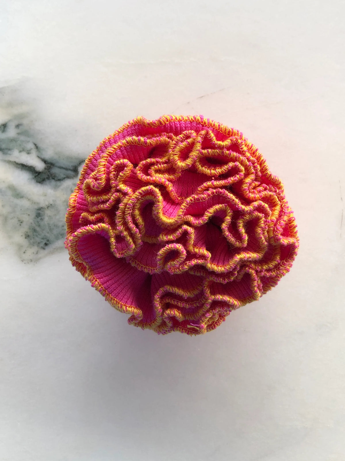 Broochella Men’s Corallo Brooch in vibrant pink fabric with contrasting golden yellow ruffled edging, photographed on a white marble background with grey-green veining.