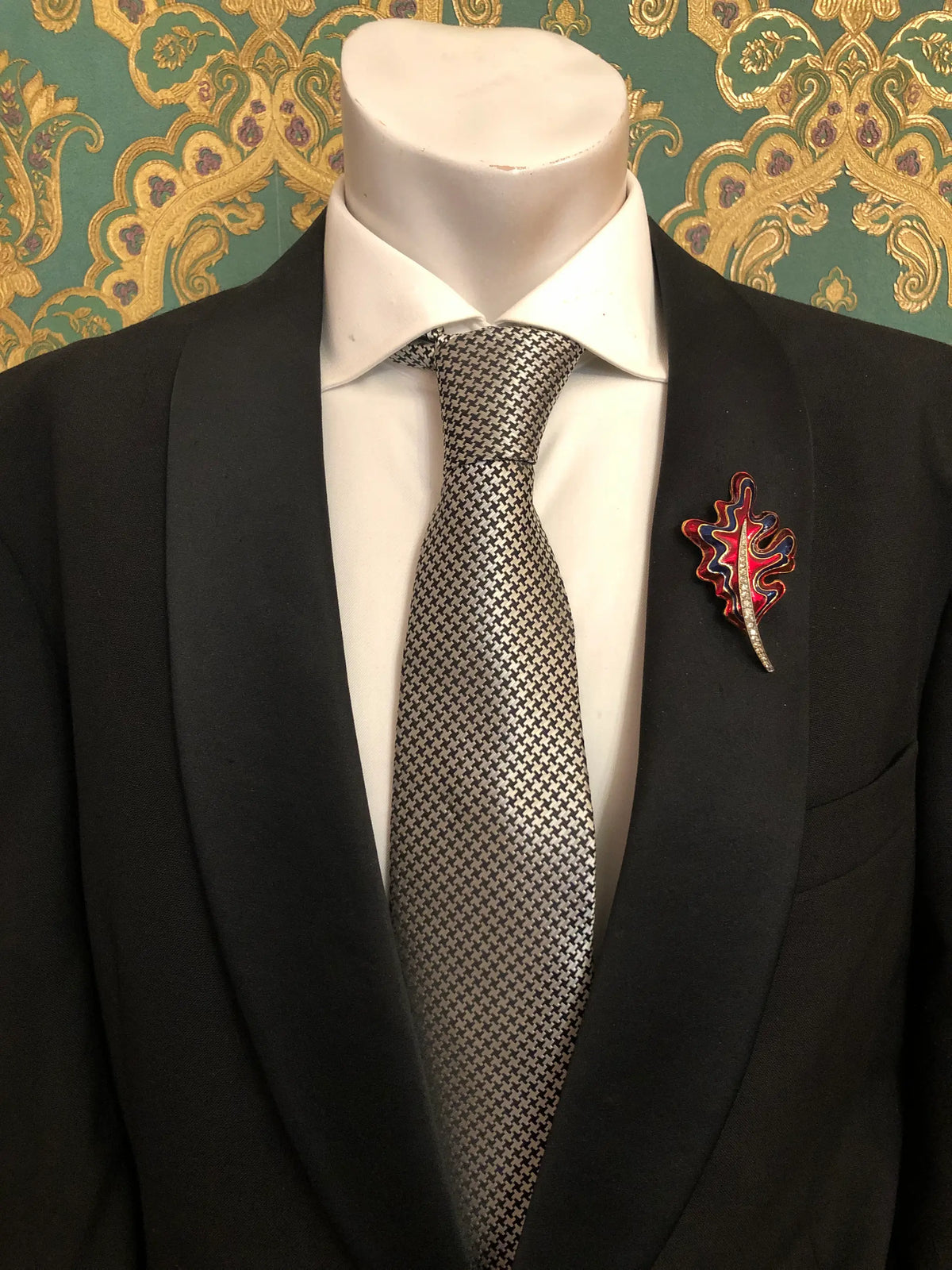 Broochella Men's Cosima vintage enamel leaf brooch featuring a sculptural oak leaf design with vibrant red and navy blue enamel, gold-tone outlines and a sparkling crystal-set stem, styled on the lapel of a black tailored suit with a white shirt and black-and-white houndstooth tie, against ornate emerald green and gold damask wallpaper.