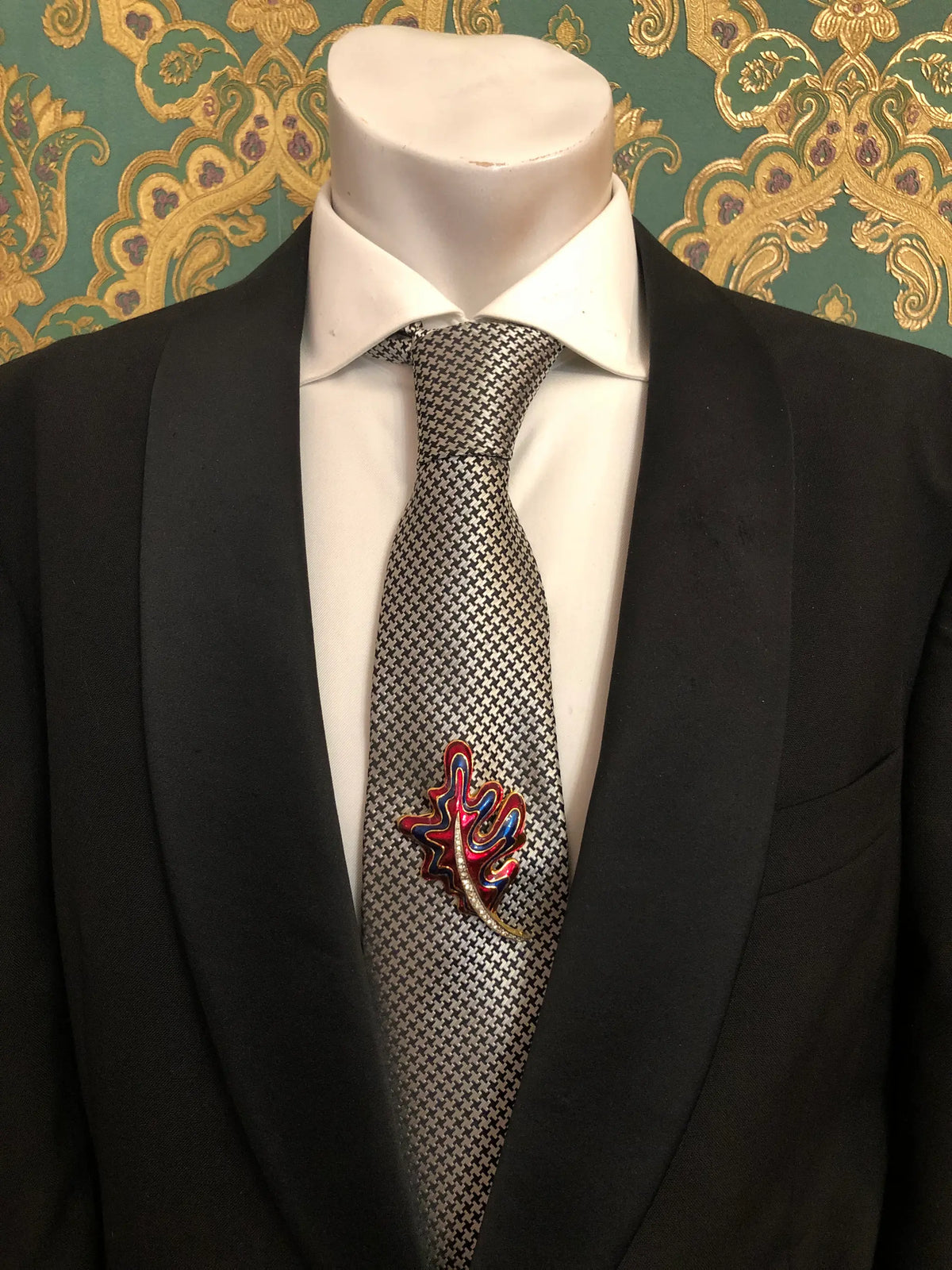Broochella Men's Cosima vintage red and navy blue enamel leaf brooch with intricate gold-tone detailing and a curved crystal-embellished stem, pinned to a black-and-white houndstooth necktie with a crisp white shirt and black tailored suit, photographed against decorative emerald green and gold damask wallpaper.