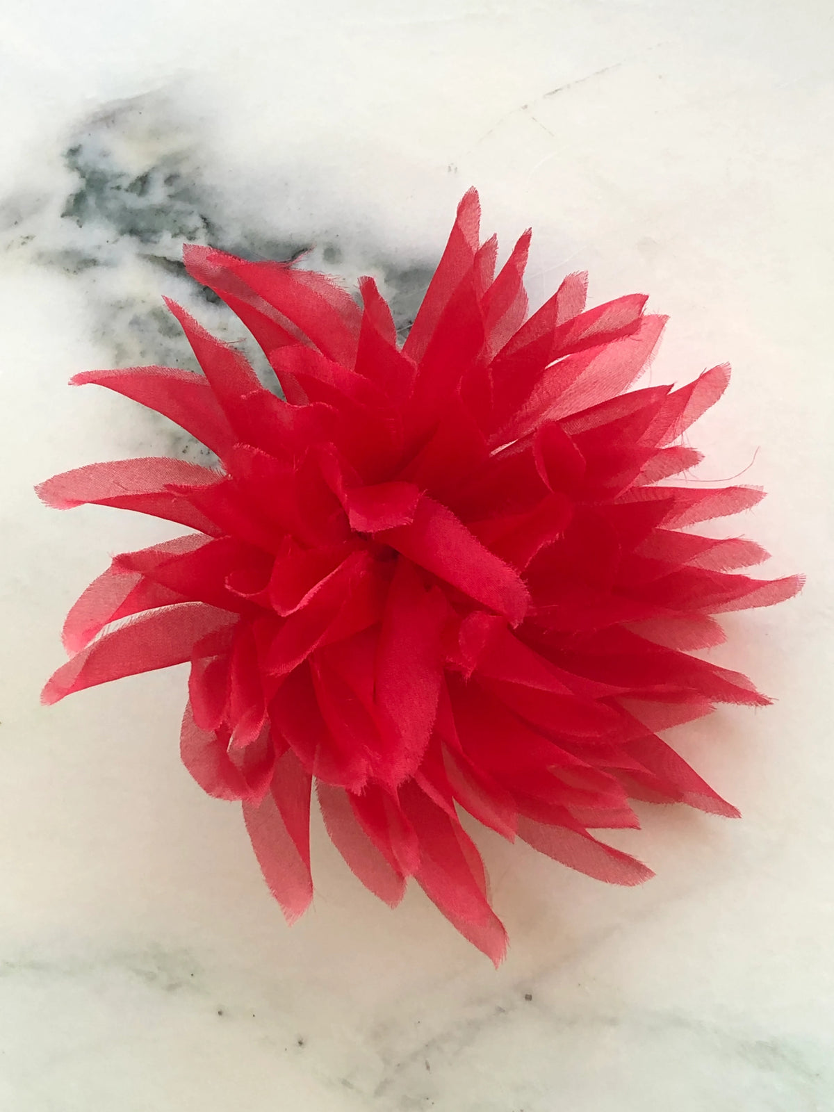 Broochella Men’s Dahlia handmade designer flower brooch in vivid red fabric, featuring abundant elongated layered petals with softly frayed edges, photographed on a white marble background with natural green veining.