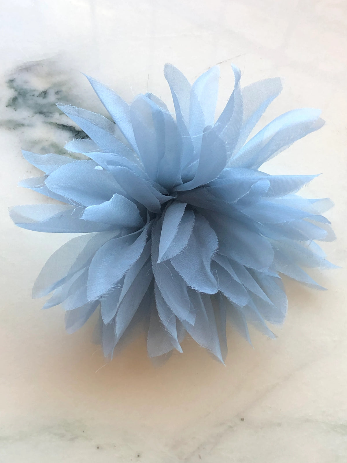 Broochella Men’s Dahlia handmade designer flower brooch in sky blue fabric, featuring abundant elongated layered petals with softly frayed edges, photographed on a white marble background with natural green veining.