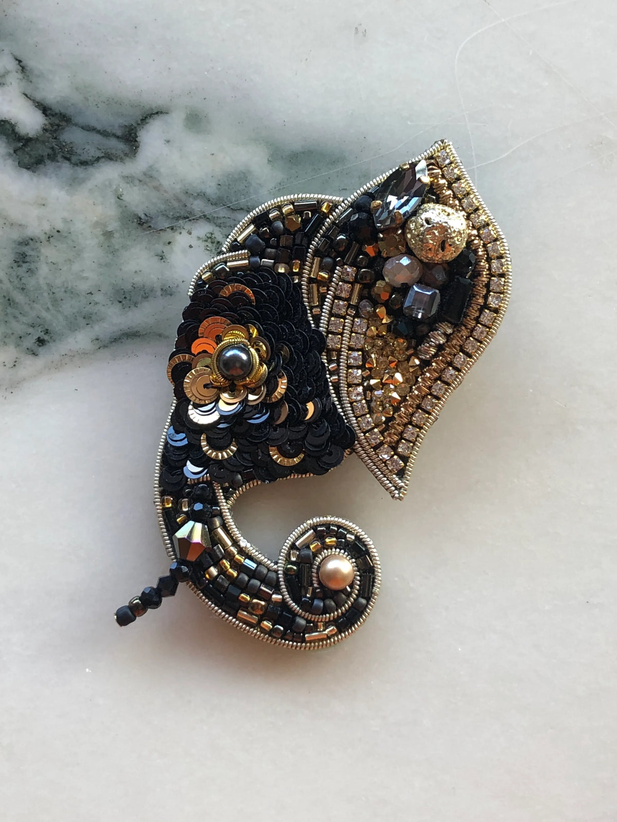 Broochella Men’s Elephant Brooch featuring intricate black, gold and bronze beadwork, sequins, crystals and pearl accents, photographed on a white marble background with grey-green veining.