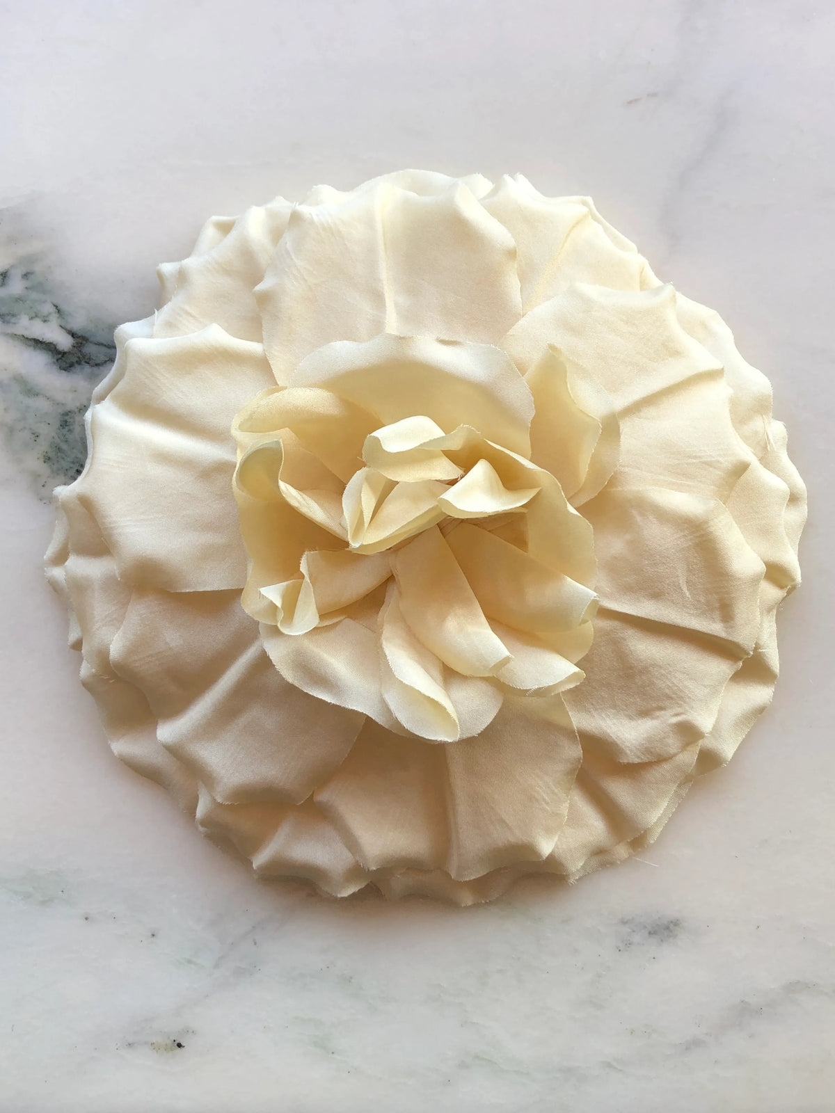 Broochella Men’s Femmina handmade designer flower brooch in soft yellow fabric, with oversized sculptural layered petals and a ruffled centre, photographed on a white marble background with natural green veining.